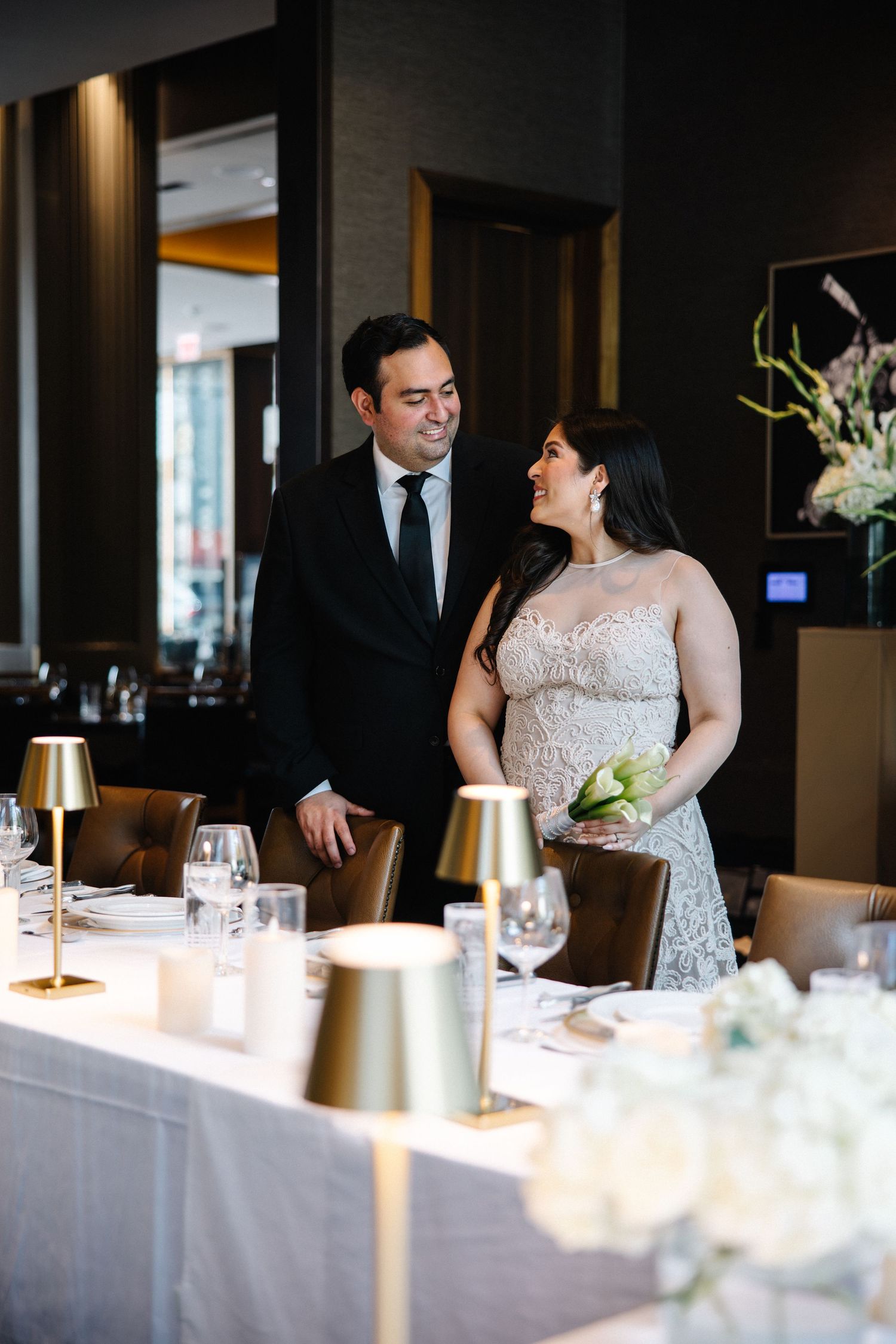 Elegant couple in wedding attire sharing a smile beside a beautifully set reception dinner table.