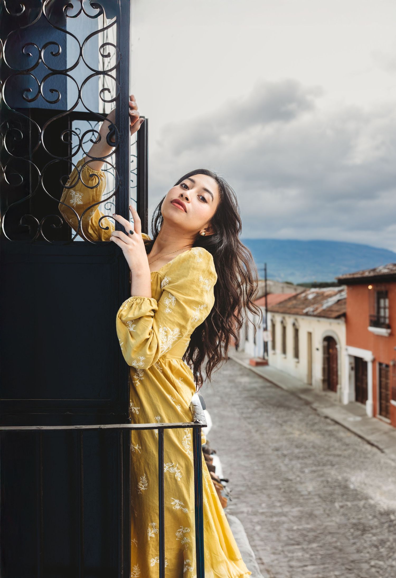 Woman in yellow floral dress leaning on ornate iron gate on scenic balcony.