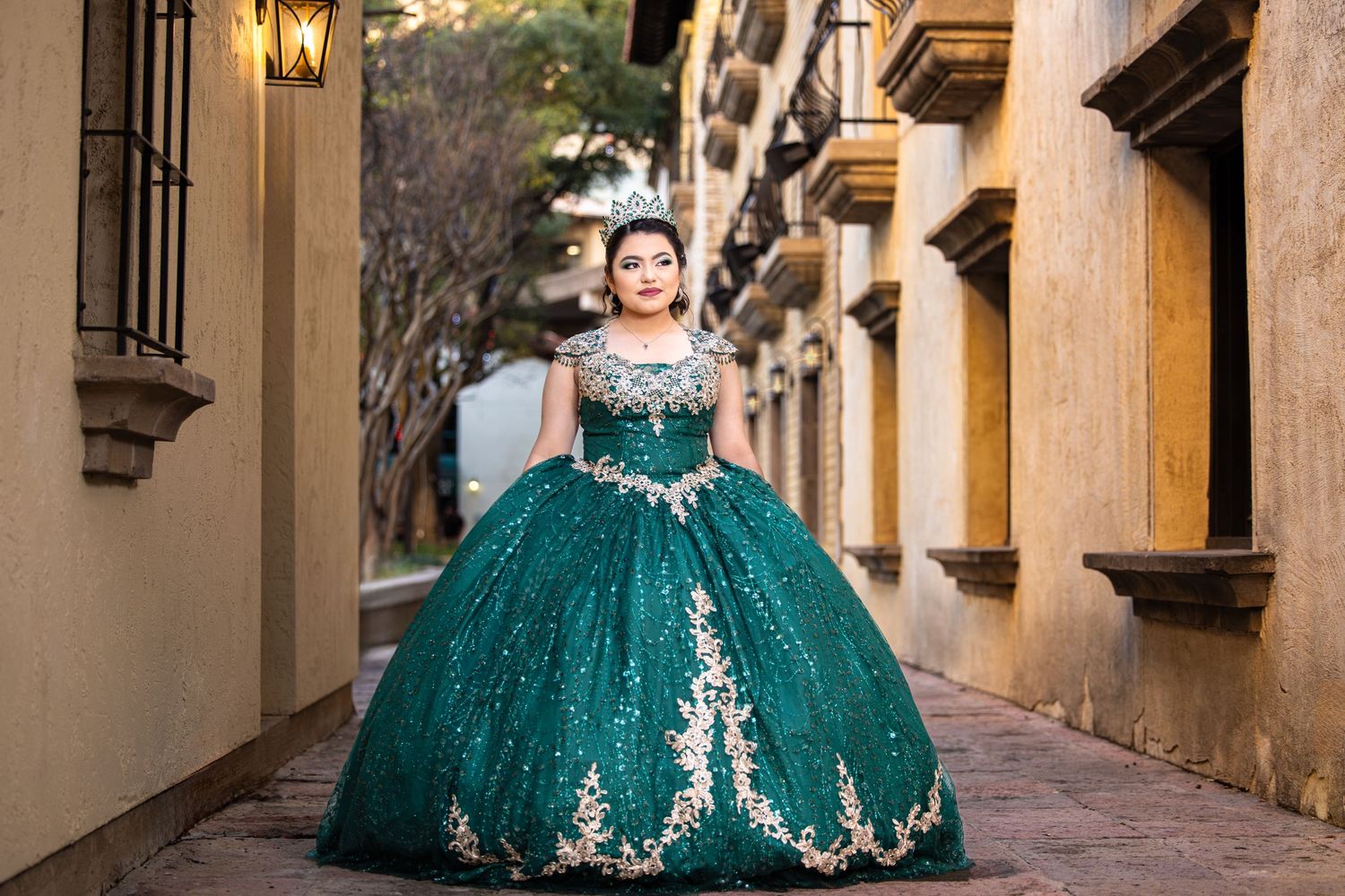 Young woman in sparkling emerald green quinceañera ball gown with gold embroidery posing on historic street.