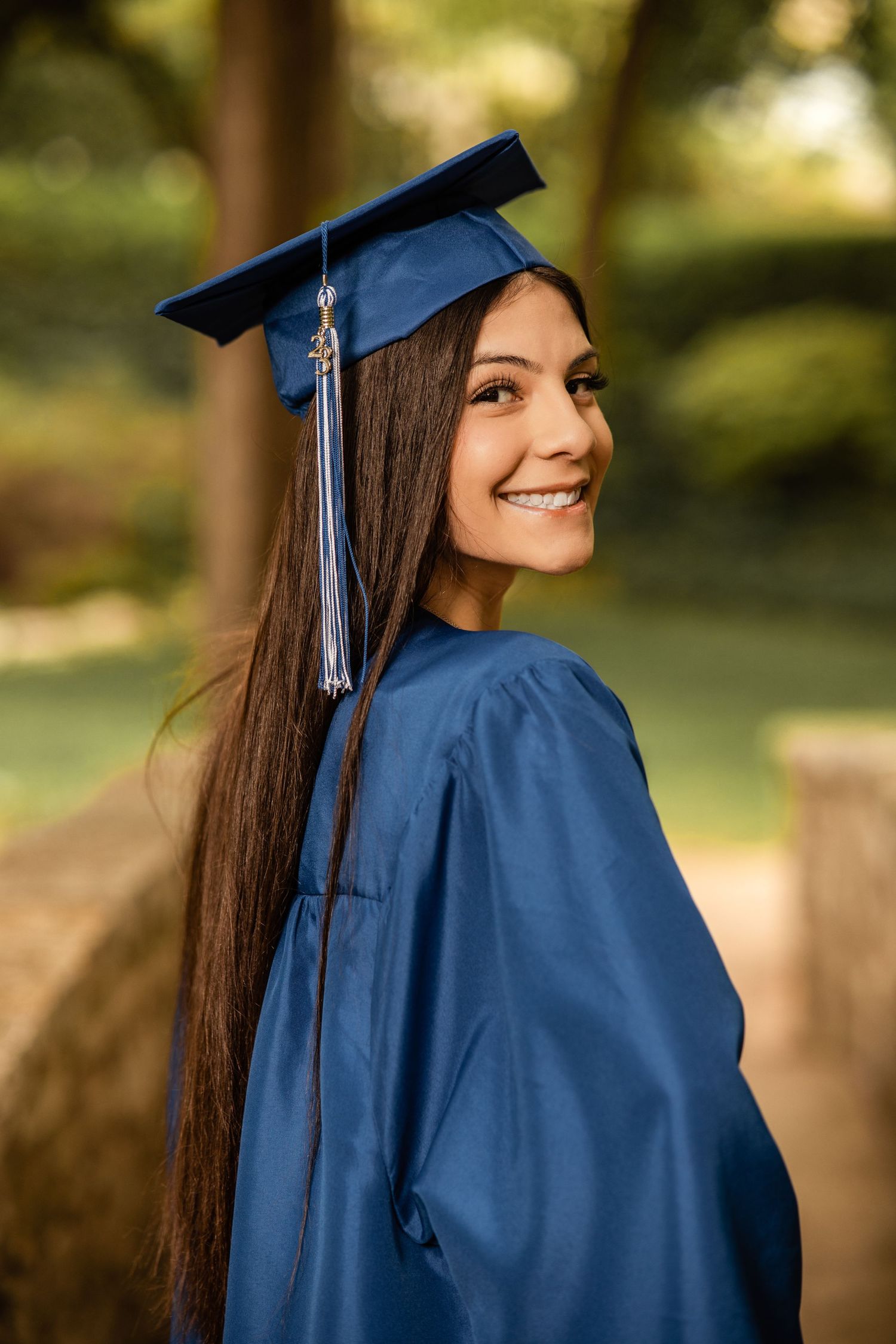 Happy female graduate in blue cap and gown smiling over shoulder in park setting.