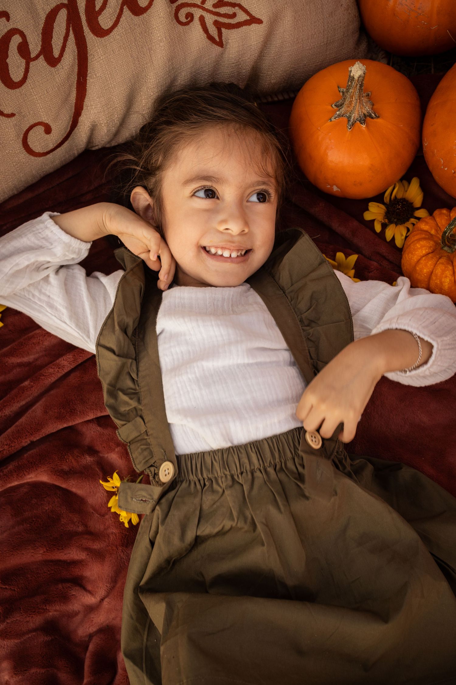 Smiling young girl lying among pumpkins and autumn decorations in a cozy fall-themed portrait.