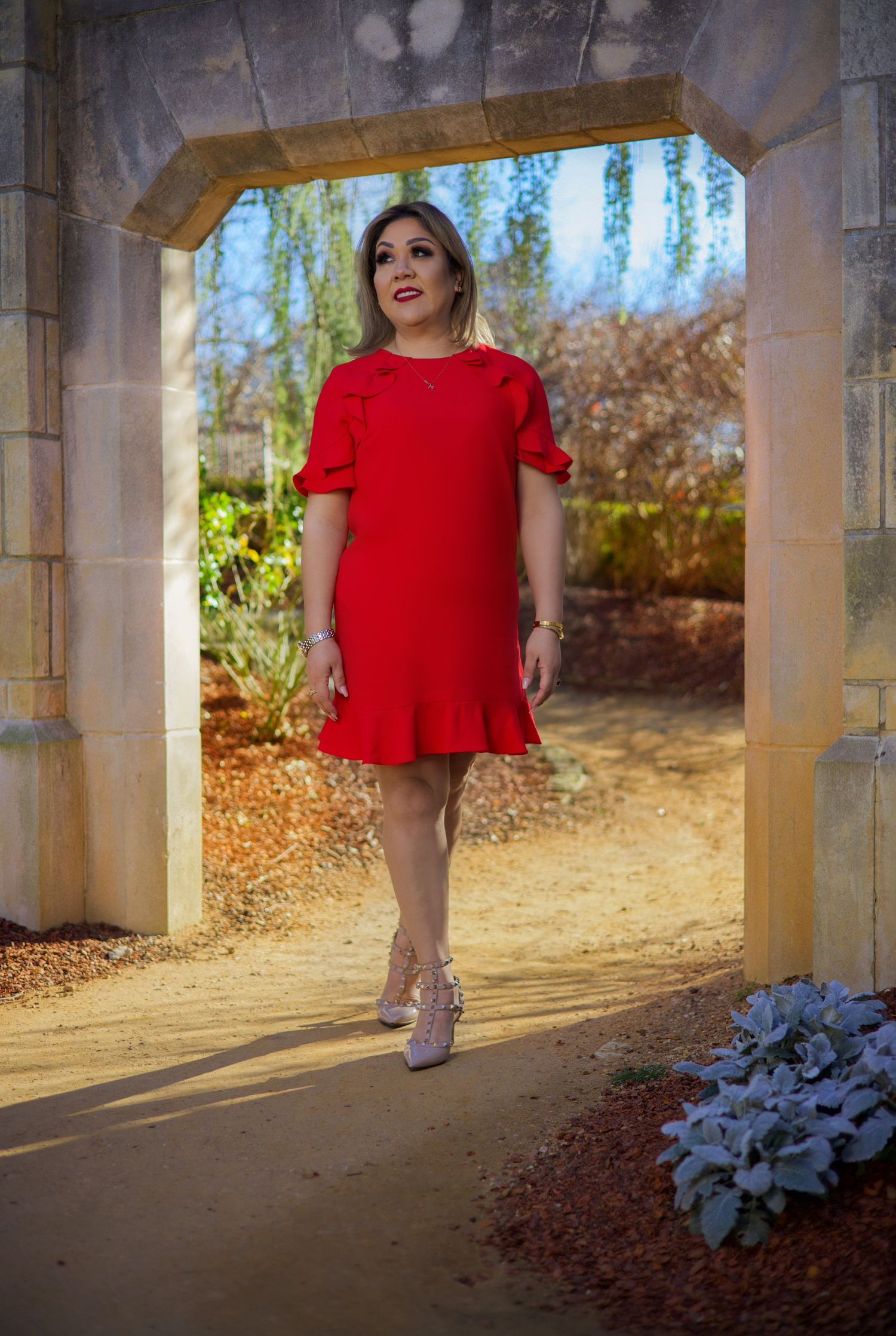 Woman in elegant red ruffle dress posing under stone archway in garden setting.