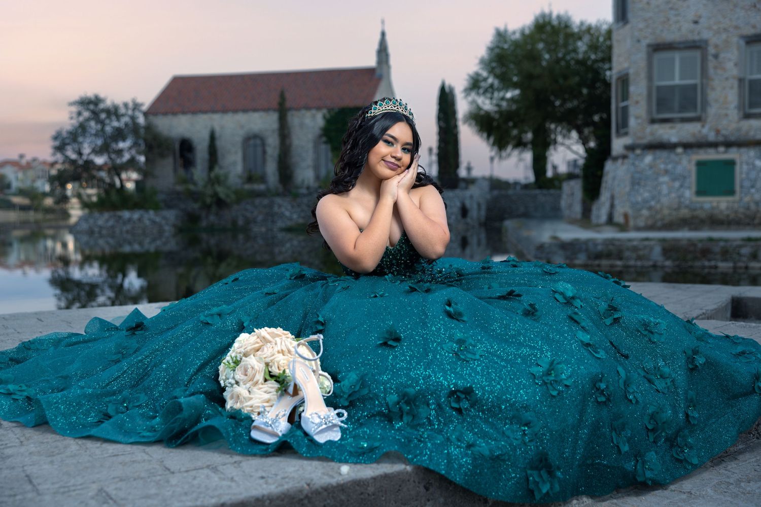 Quinceañera girl in teal ballgown with floral details sits gracefully holding white bouquet near stone chapel.