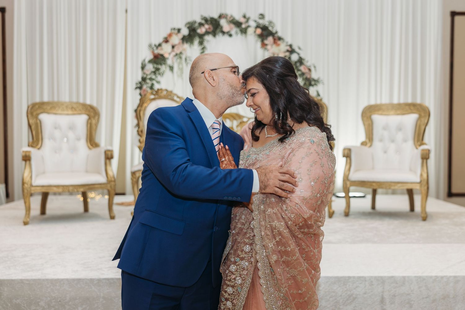 Groom kisses bride wearing beaded pink saree before elegant floral arch in wedding venue.