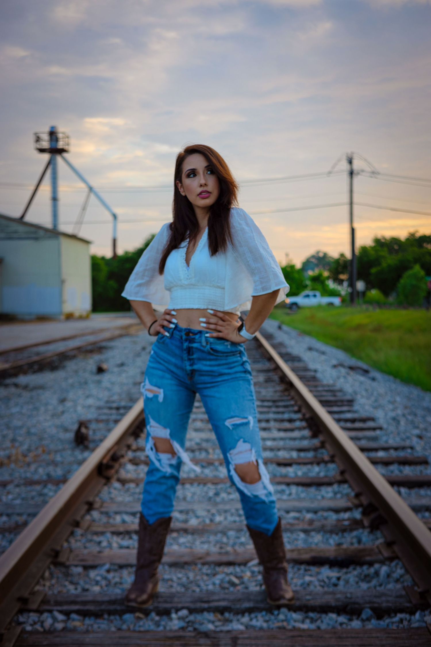 Woman in white crop top and ripped jeans posing on railroad tracks at dusk.