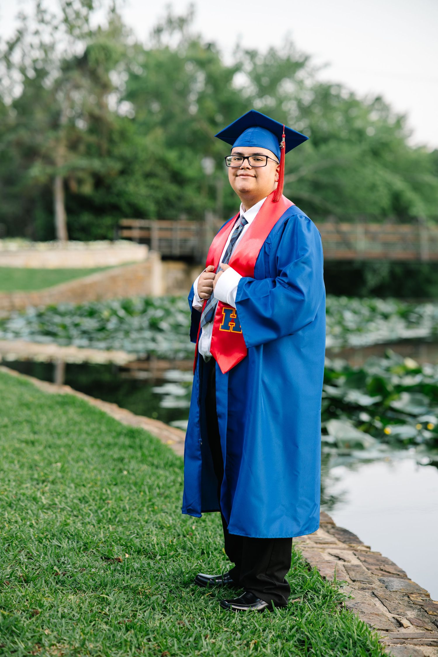 Graduate in blue cap and gown standing proudly beside a serene pond in a park.