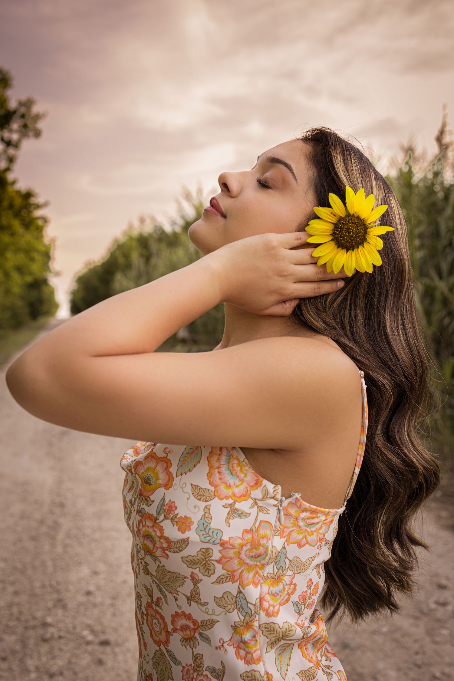 Young woman with wavy hair holding a sunflower against her cheek on a rural dirt road.