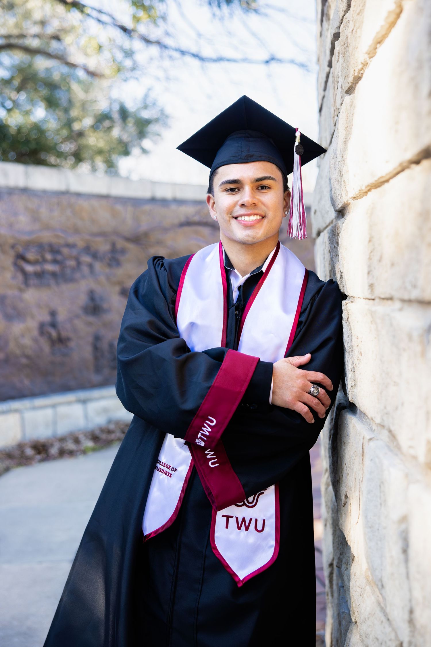 Smiling male graduate in black cap and gown with TWU stoles leaning against stone wall.