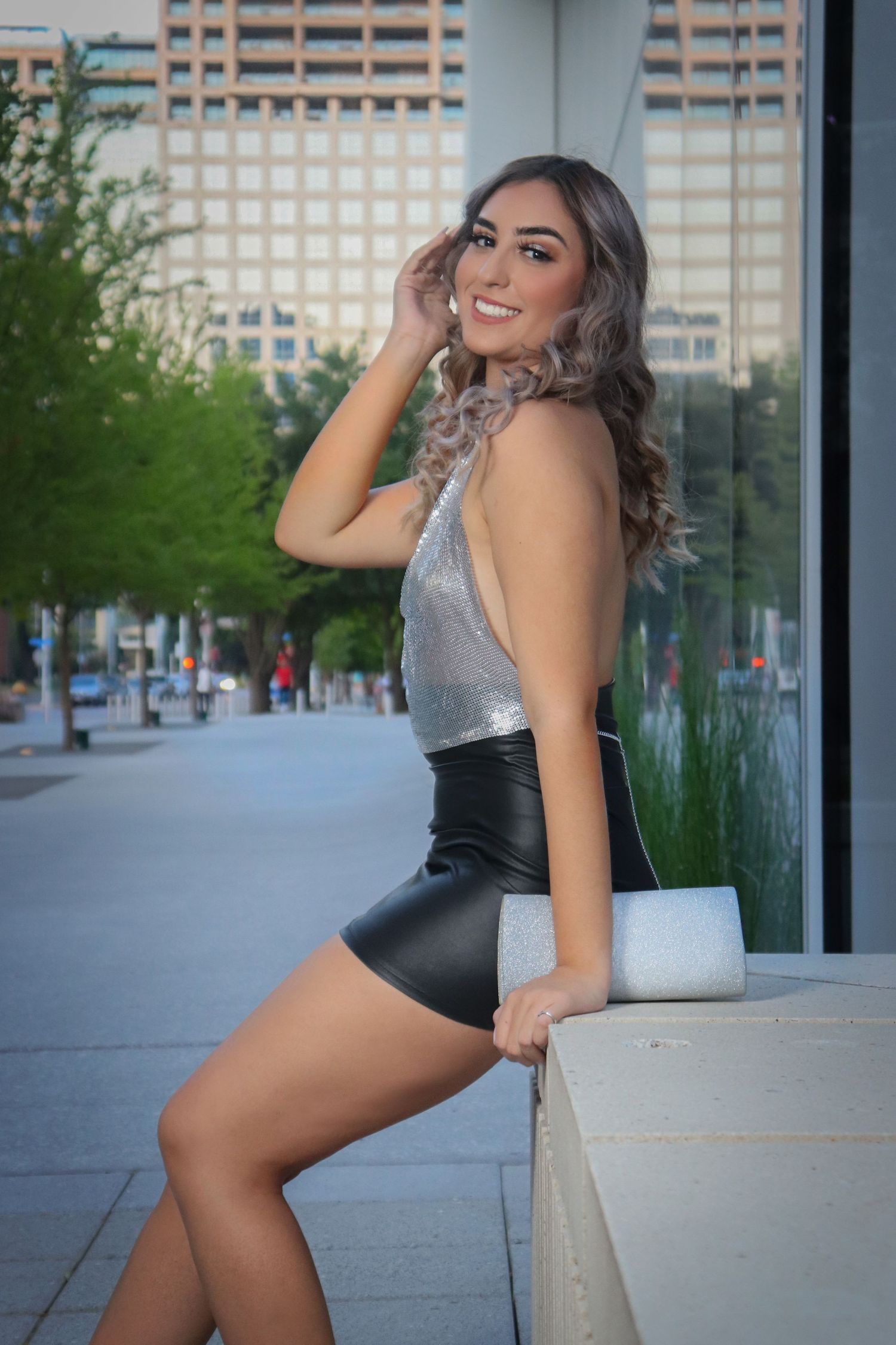 Young woman in silver top and black leather mini skirt posing outdoors in urban setting.