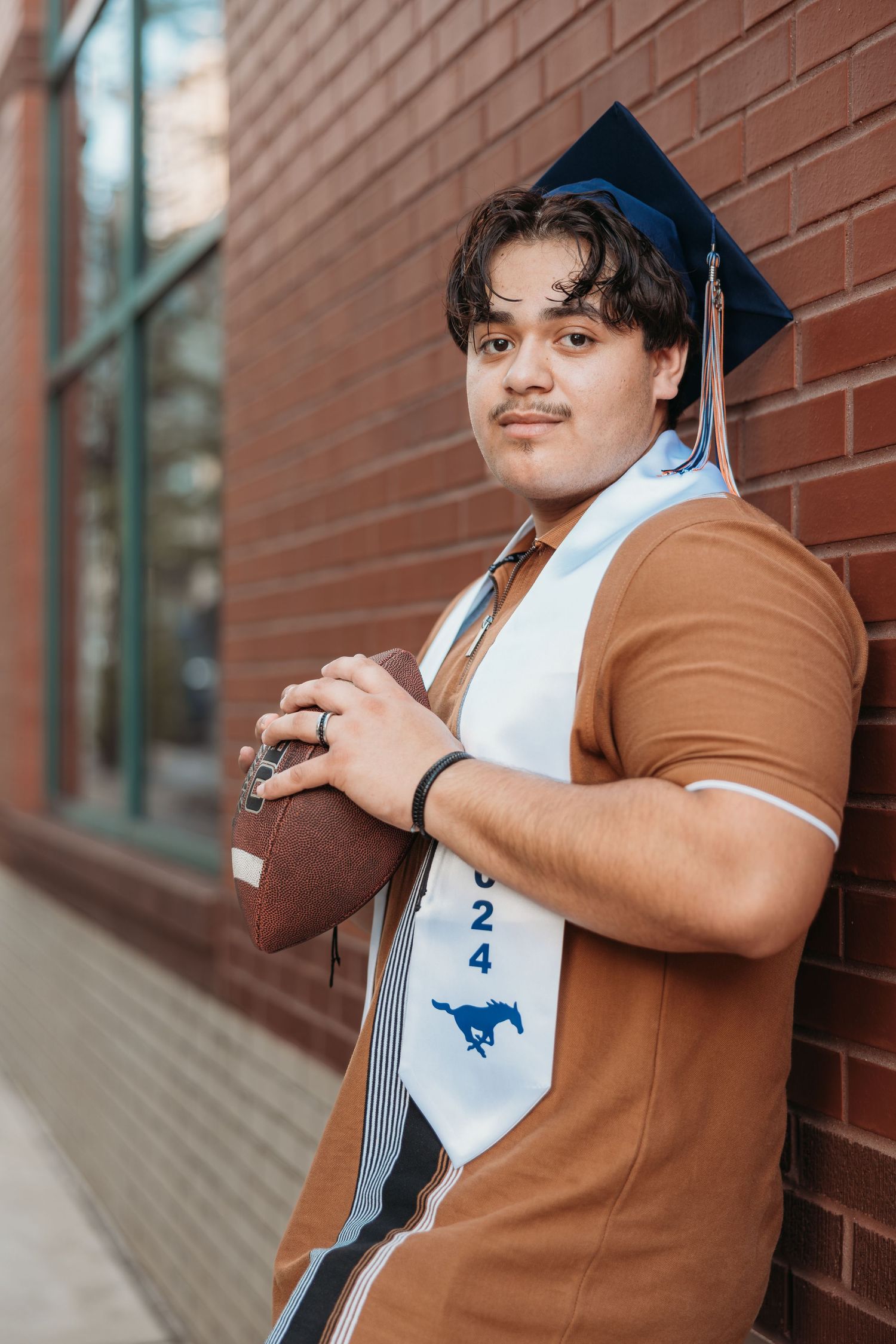 Young man in graduation cap and stole holding football against brick wall for senior photos.