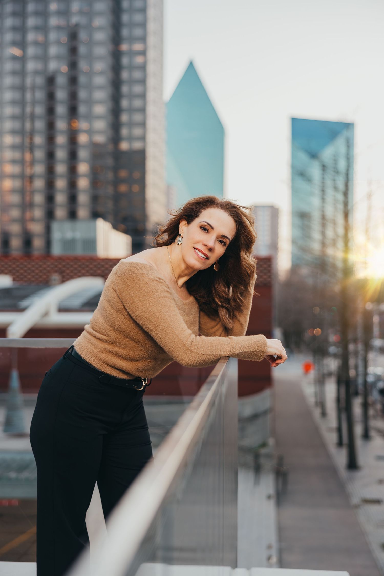 Woman in tan sweater leaning on railing with Dallas skyline at golden hour.