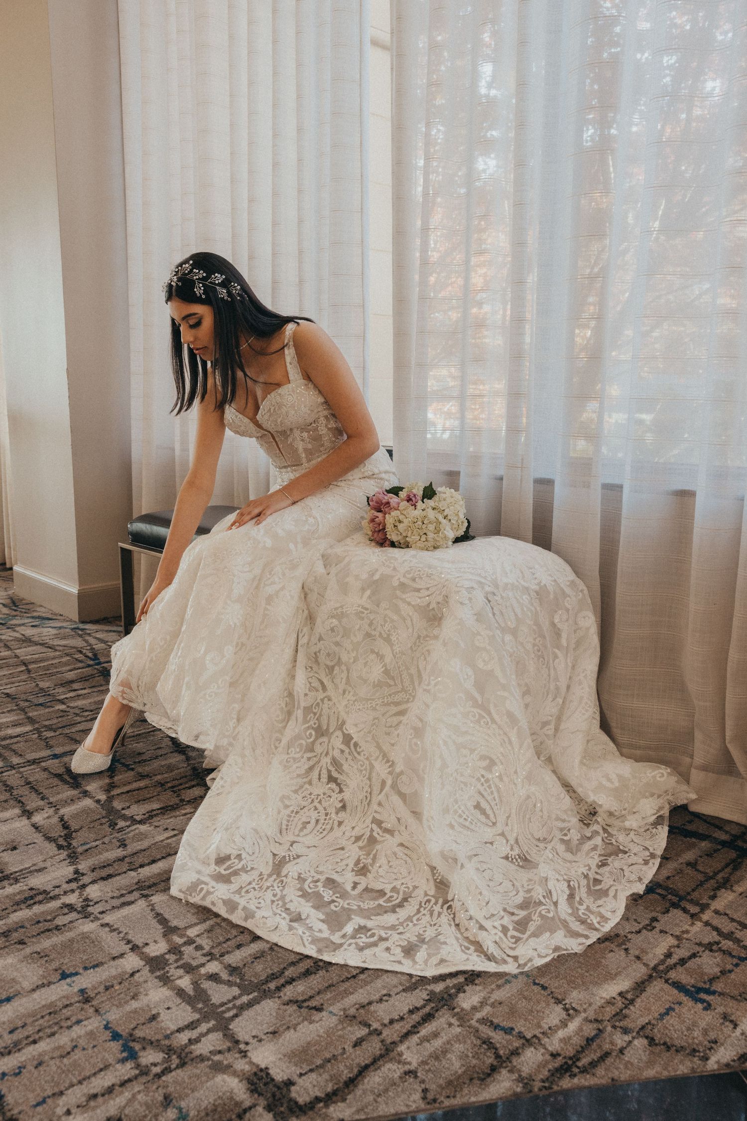 Bride in floral lace wedding gown sits by sheer curtained window wearing floral hair crown.