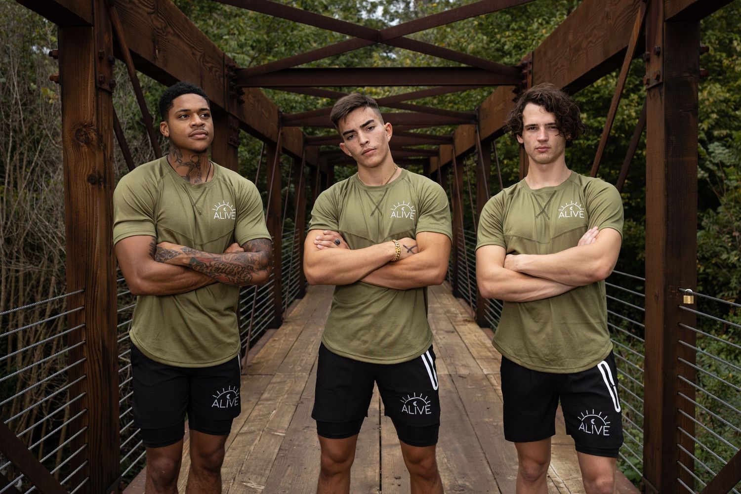 Three athletic men in matching olive green shirts posing on wooden bridge.