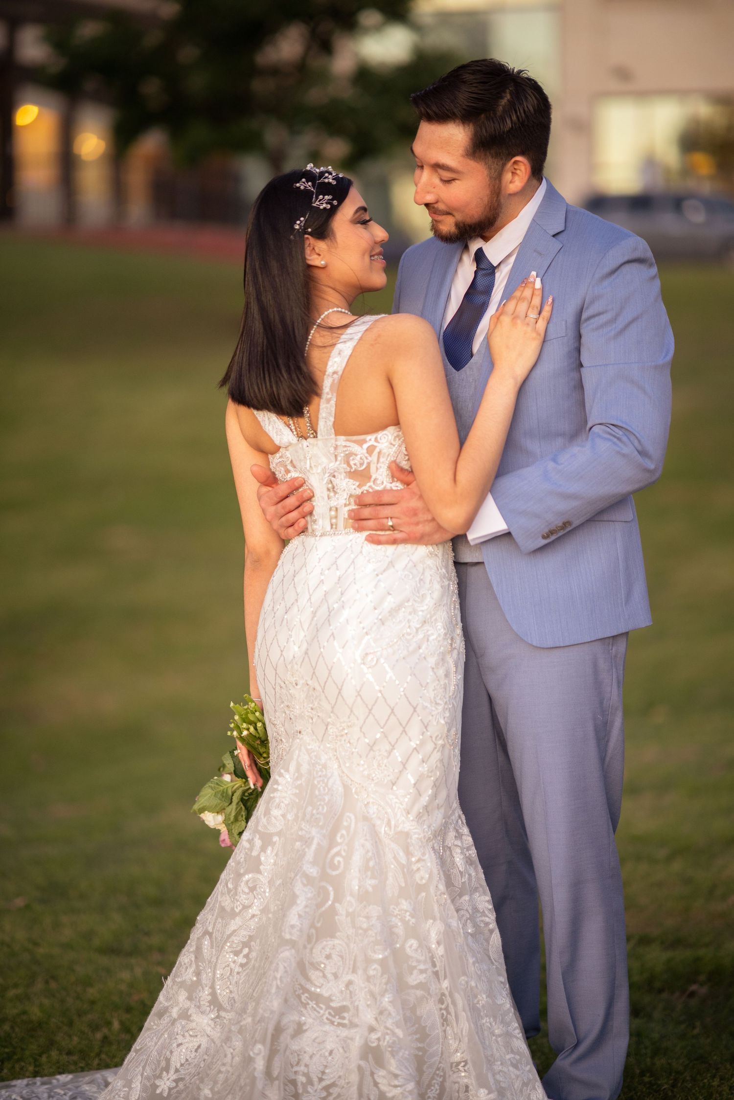 Bride and groom in blue suit share romantic embrace on green lawn at golden hour.