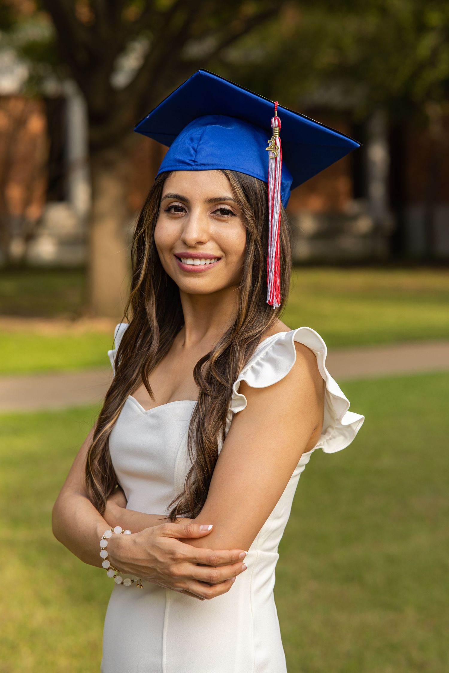 Smiling young woman in blue graduation cap and white dress with arms crossed outdoors.