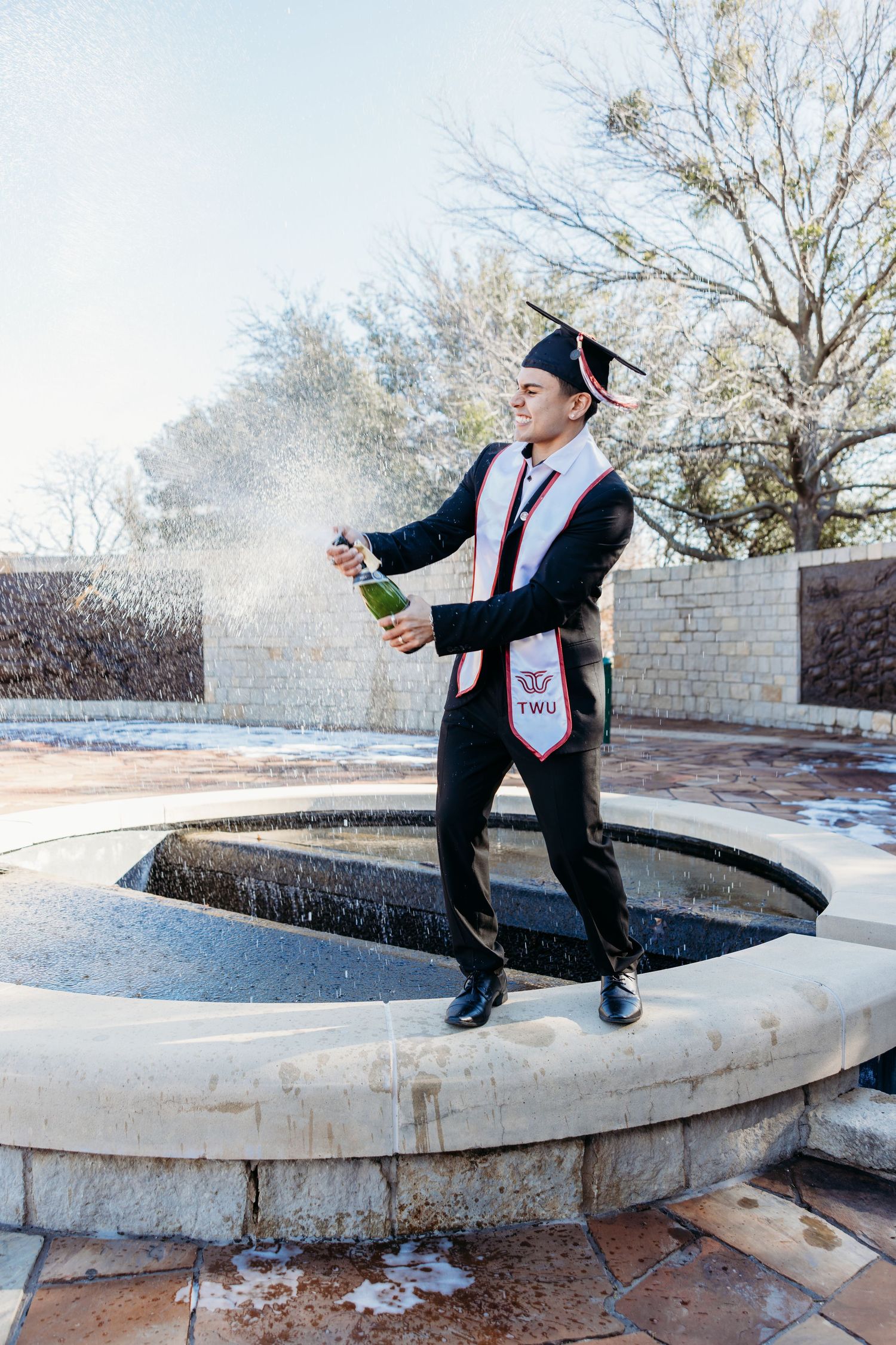 Excited TWU graduate popping champagne bottle while standing on fountain ledge outdoors.