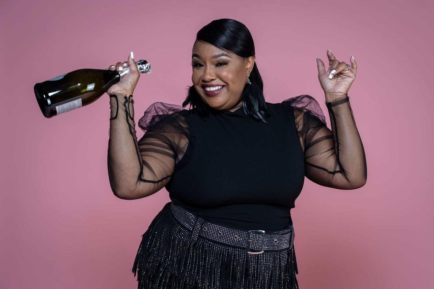 Joyful plus-size woman in black outfit holding a champagne bottle on pink background.