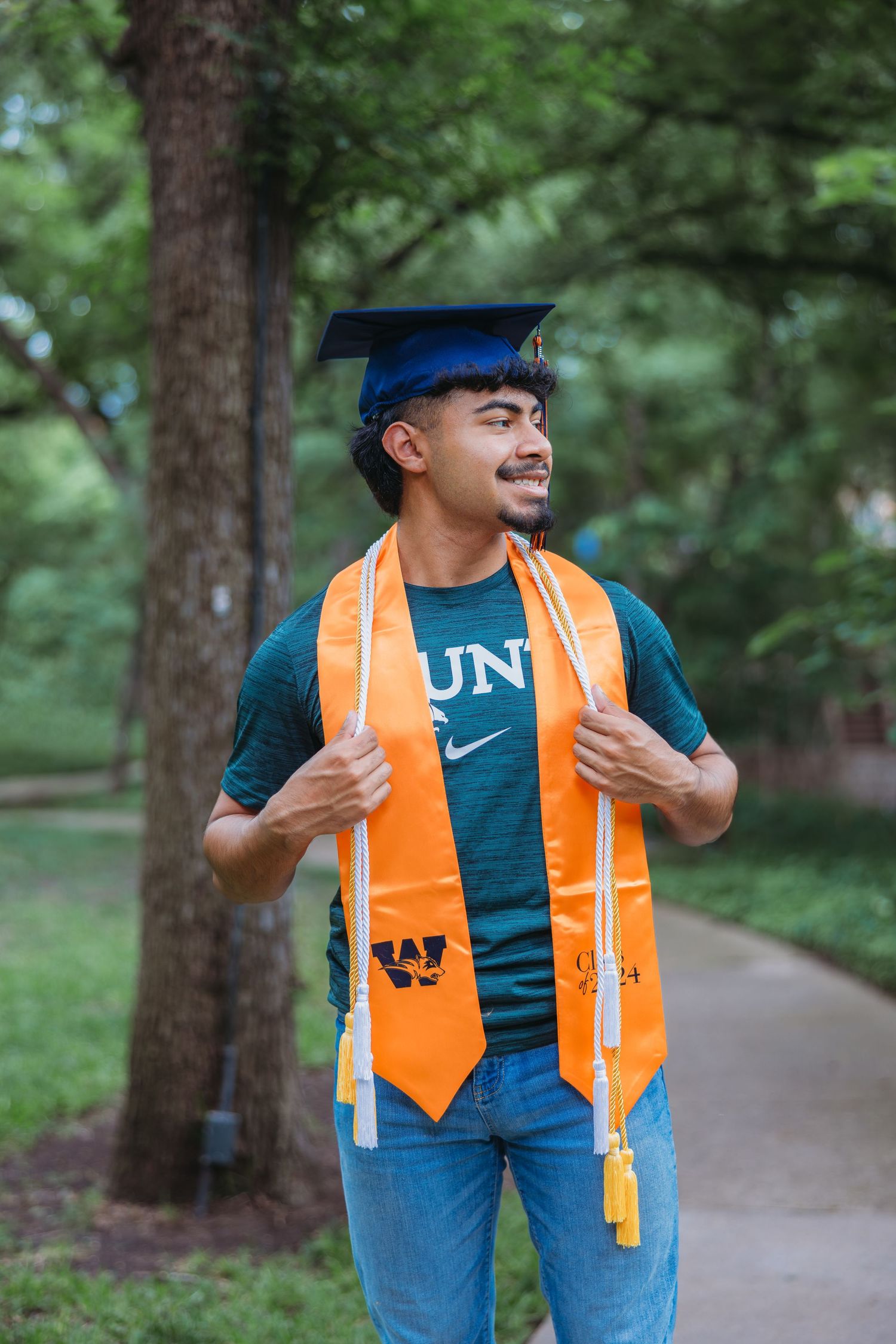 Smiling male graduate in blue cap with orange honor stole walking through tree-lined campus path.