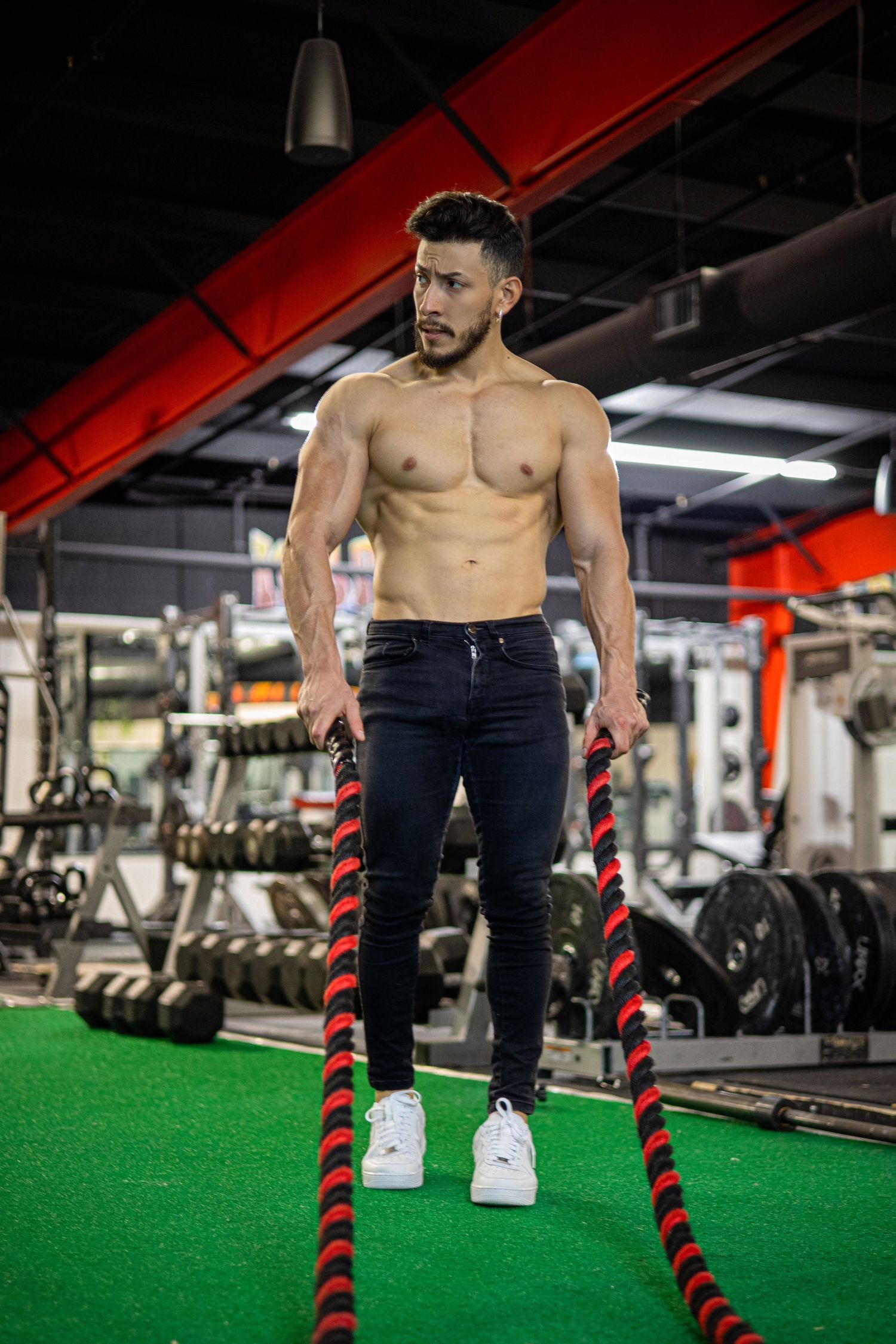 Muscular shirtless man holding battle ropes during intense workout in modern gym.