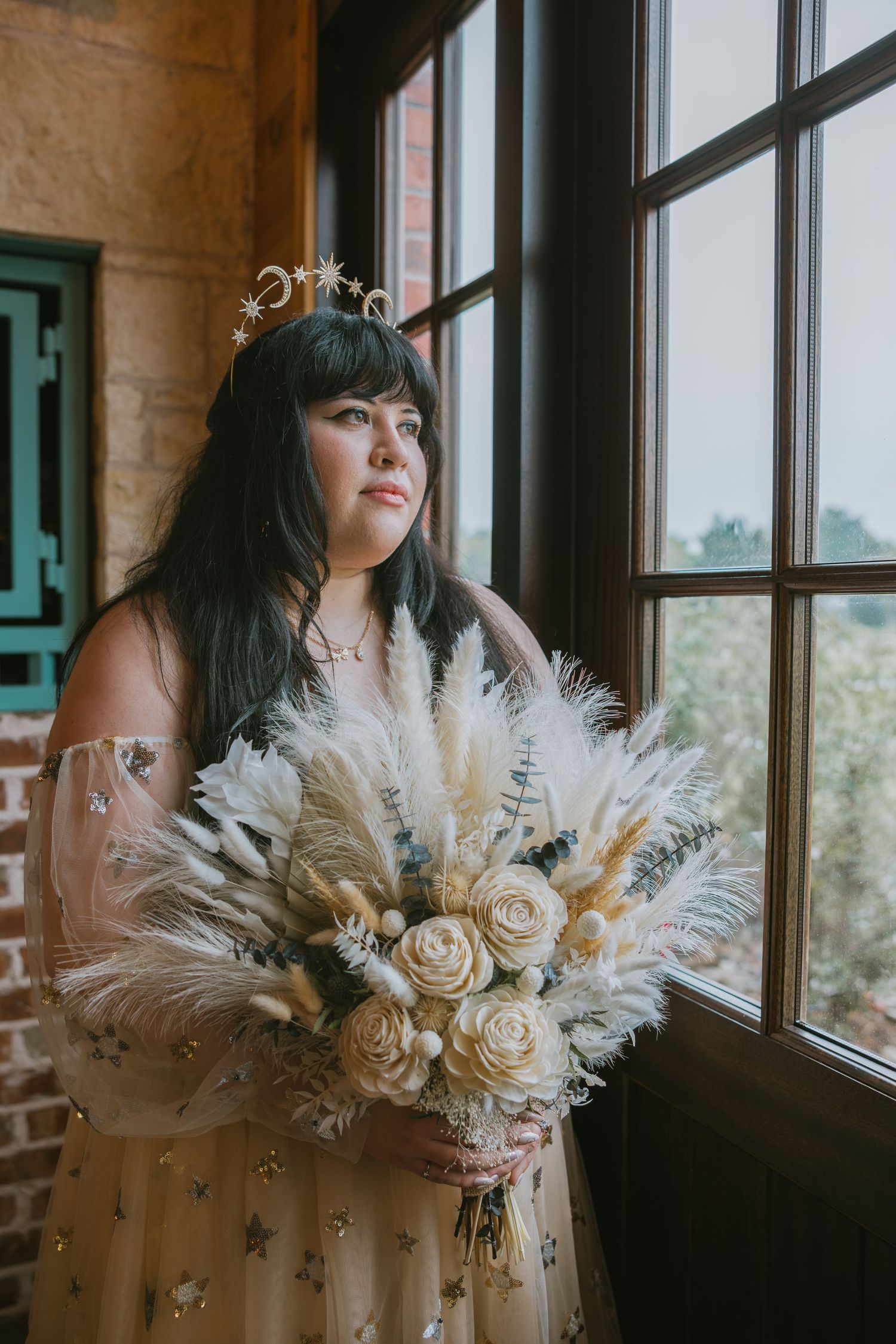 Bride with dark hair wearing a celestial crown holding a boho pampas grass bouquet by a large window.