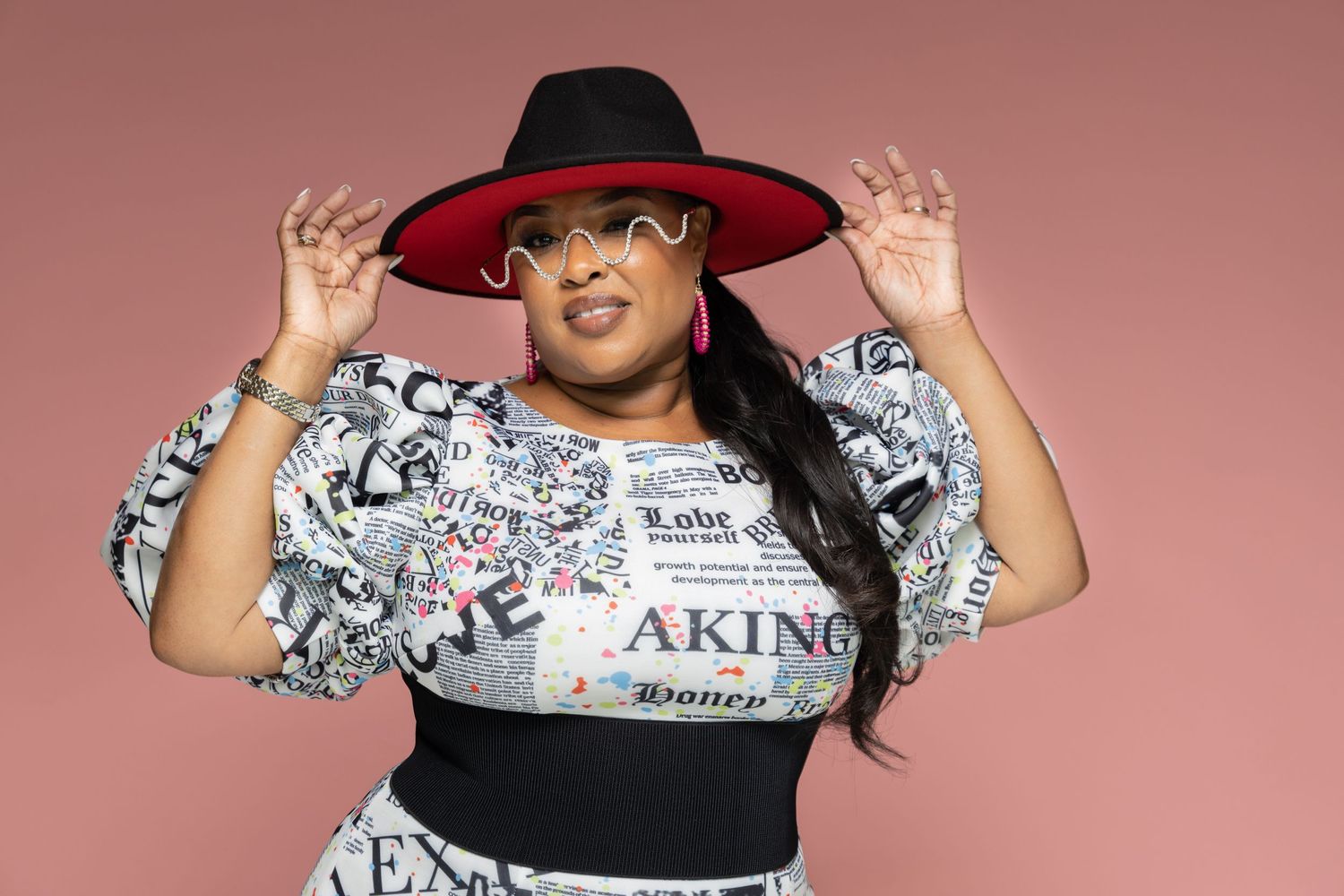 Plus-size woman posing confidently in a graphic print top and black hat on pink background.