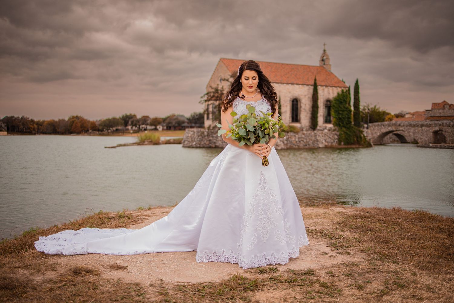 Bride in white ball gown with long train holds bouquet near lakeside chapel under dramatic cloudy sky.