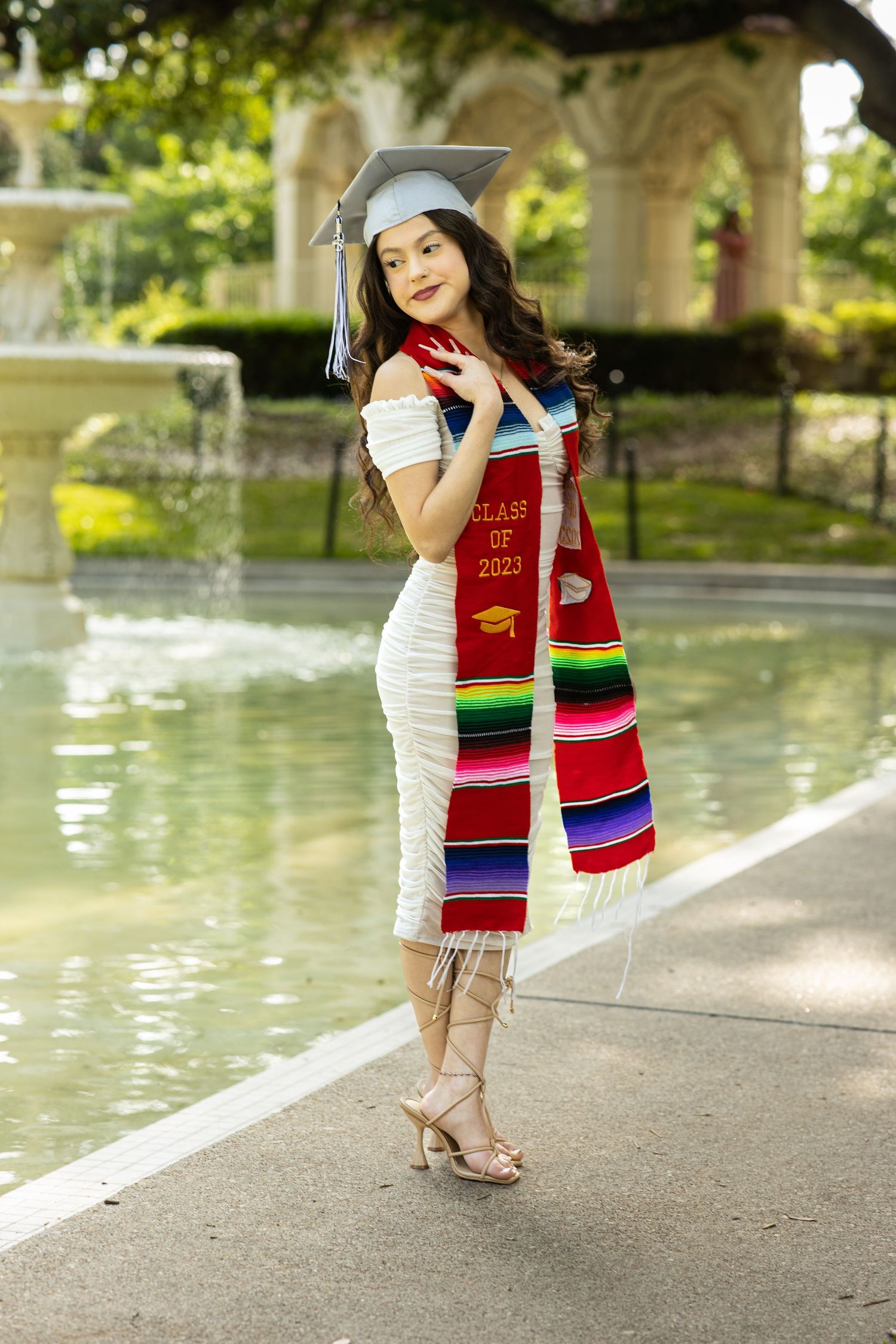 Young woman in white dress with Class of 2023 graduation stole posing by fountain.