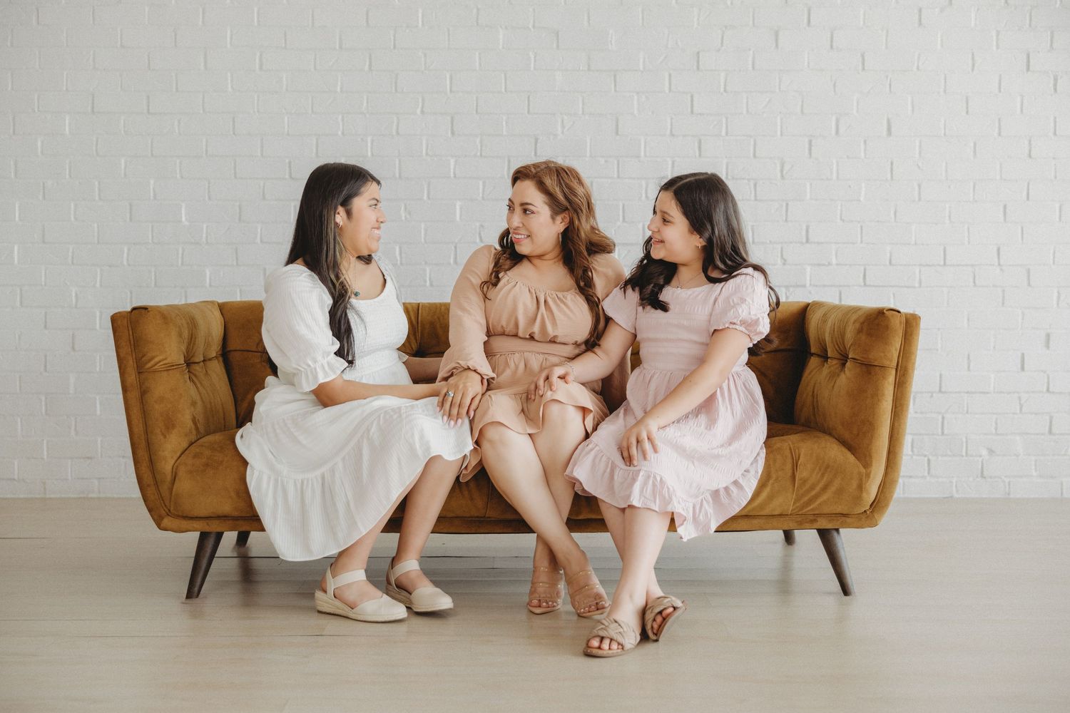 Three women sitting together on a brown velvet sofa in a bright white room.