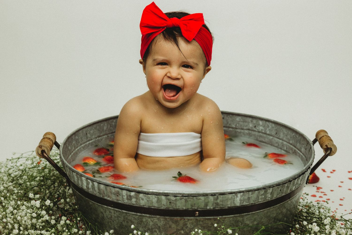 Happy laughing baby girl with red bow headband splashing in a decorative milk bath with strawberries.