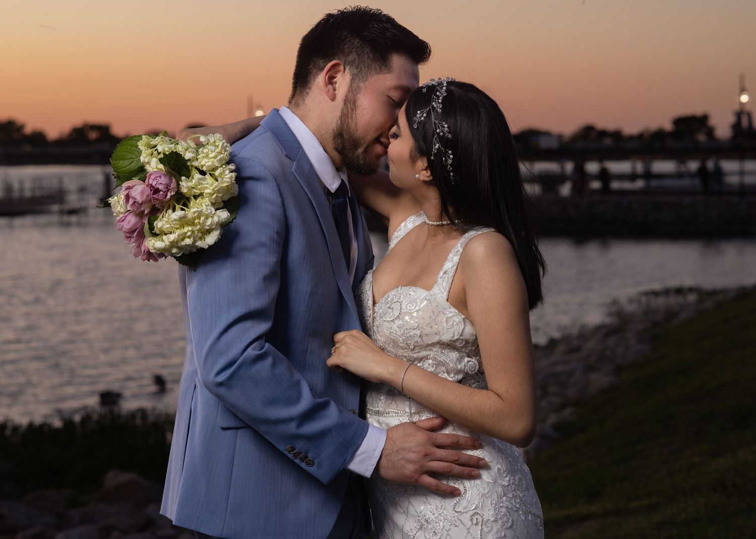 Bride and groom sharing romantic kiss near waterfront at sunset, she wears lace gown with floral headpiece.