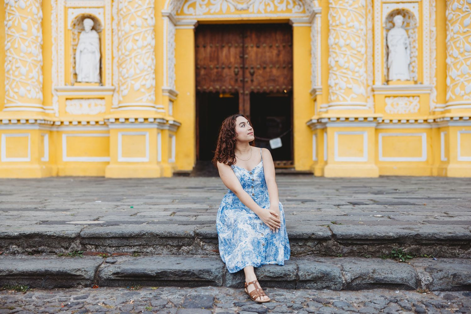Young woman in blue floral dress sitting on steps before a vibrant yellow colonial church.