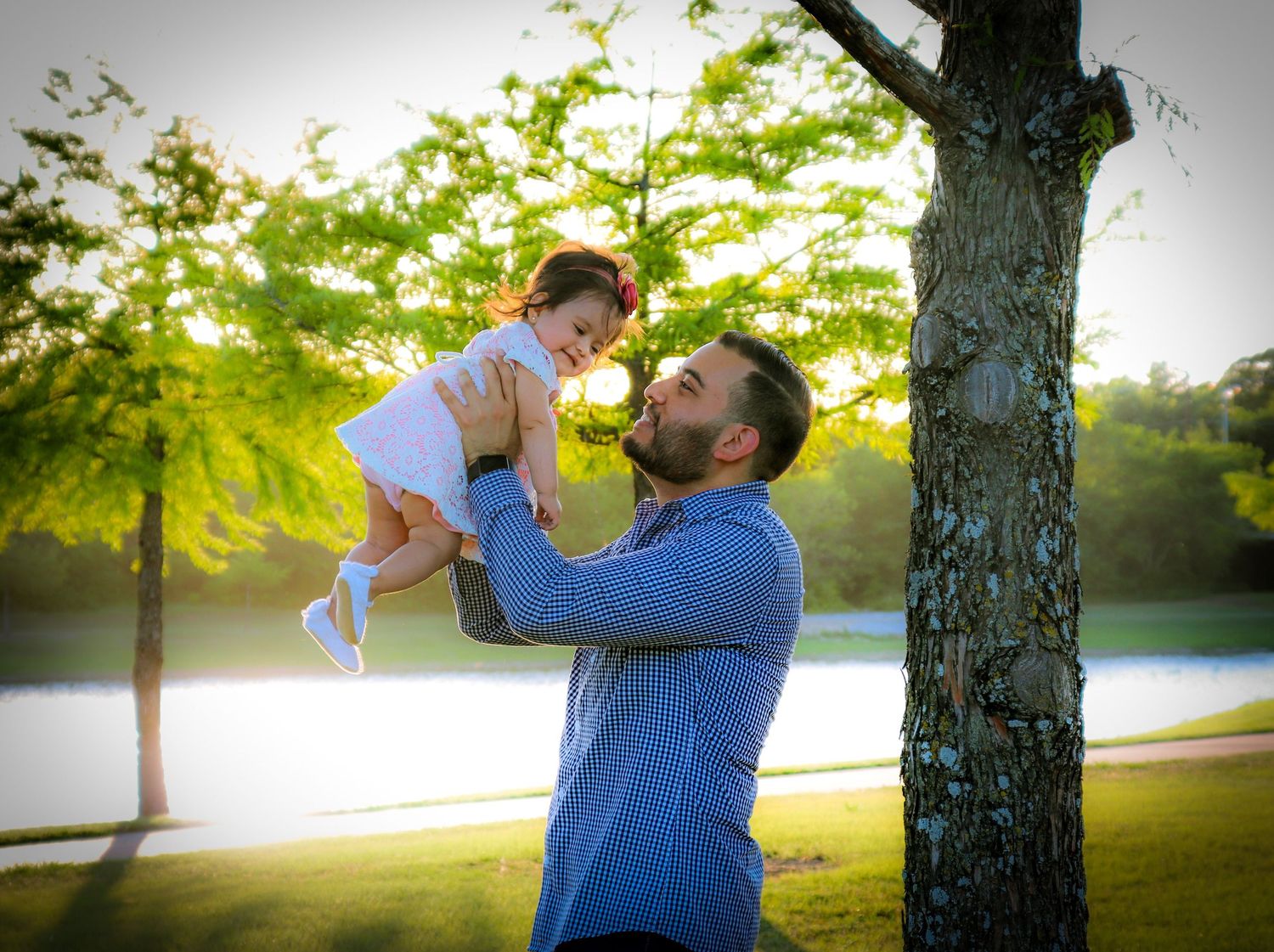 Bearded father joyfully lifting smiling toddler daughter in the air outdoors near a tree and lake.
