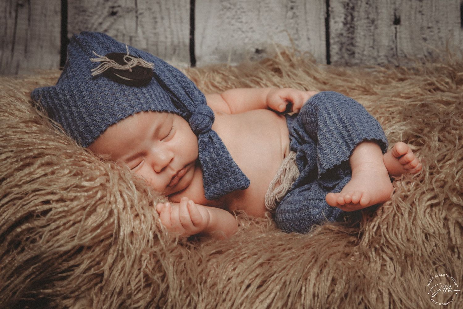 Sleeping newborn baby wearing a blue knit hat and pants on a fluffy brown fur blanket.