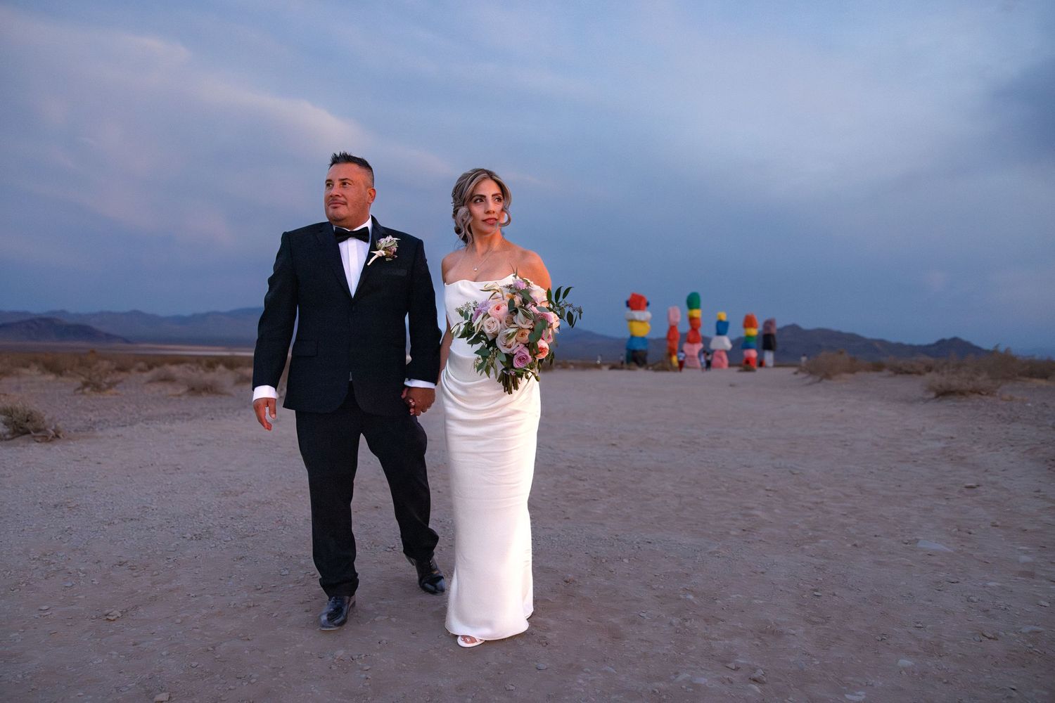Bride and groom walking in desert landscape at dusk with colorful Seven Magic Mountains art installation in background.
