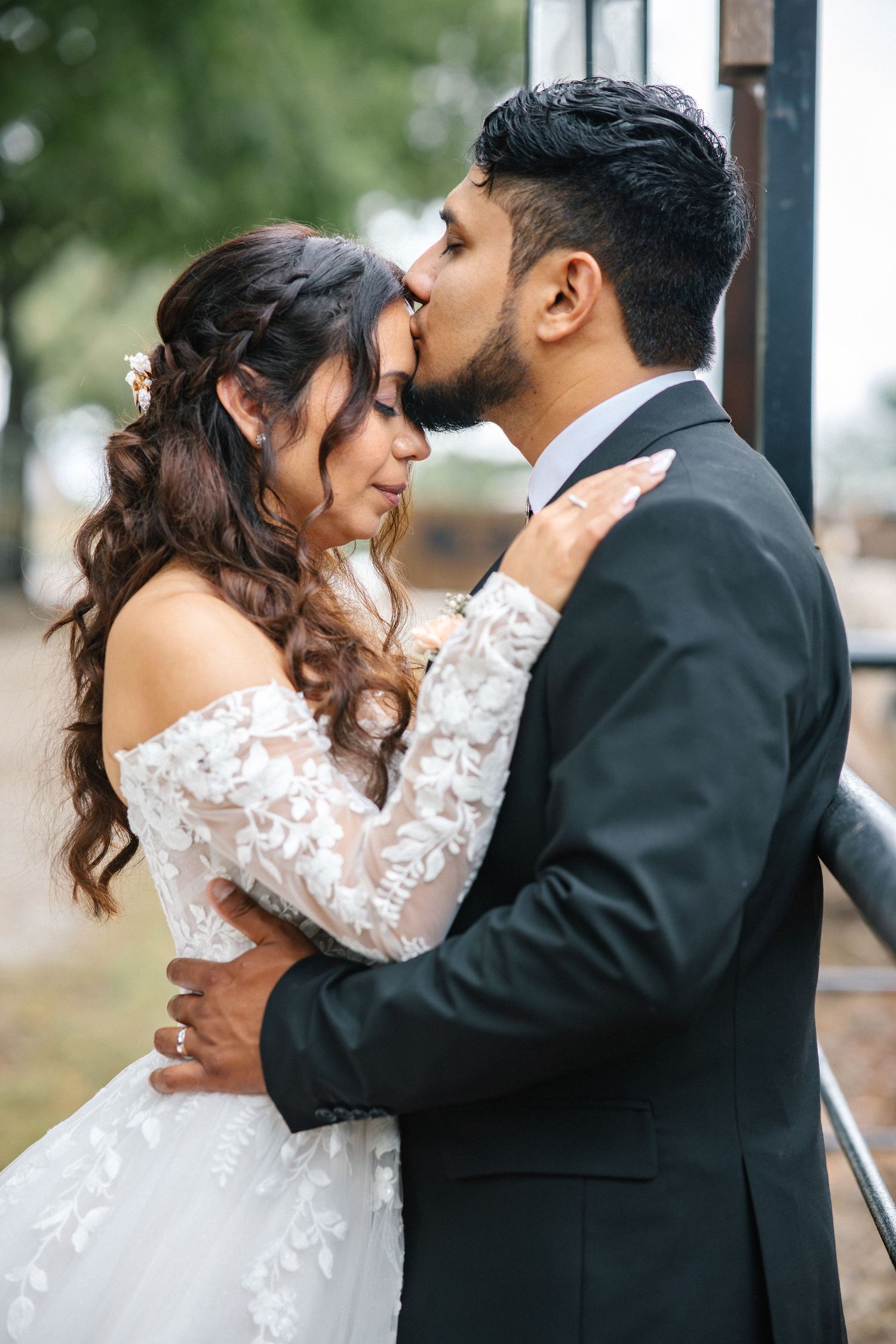 Groom in dark suit kisses bride on forehead as she embraces him in lace wedding gown.