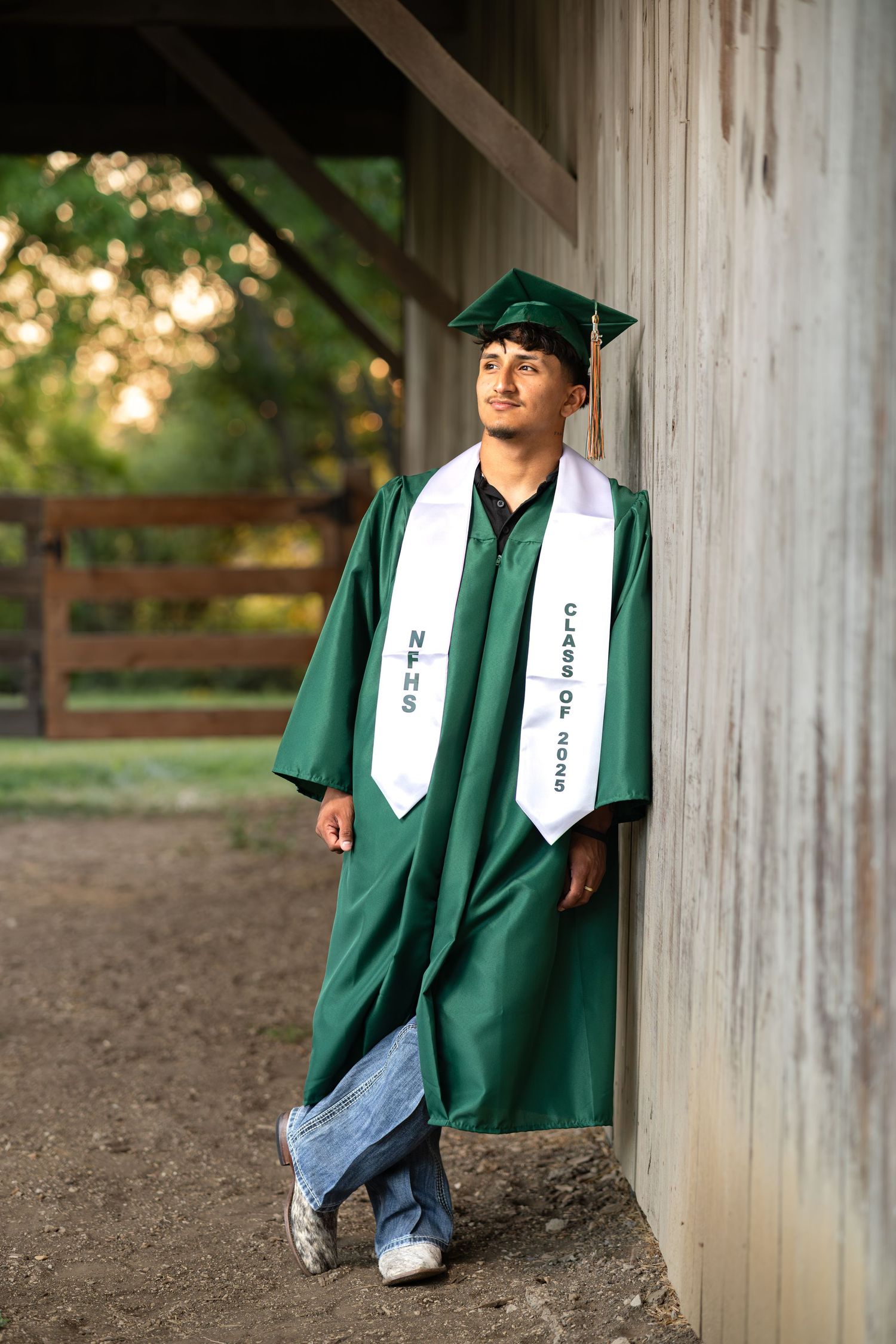 Male high school graduate in green cap and gown leaning against barn wall outdoors.