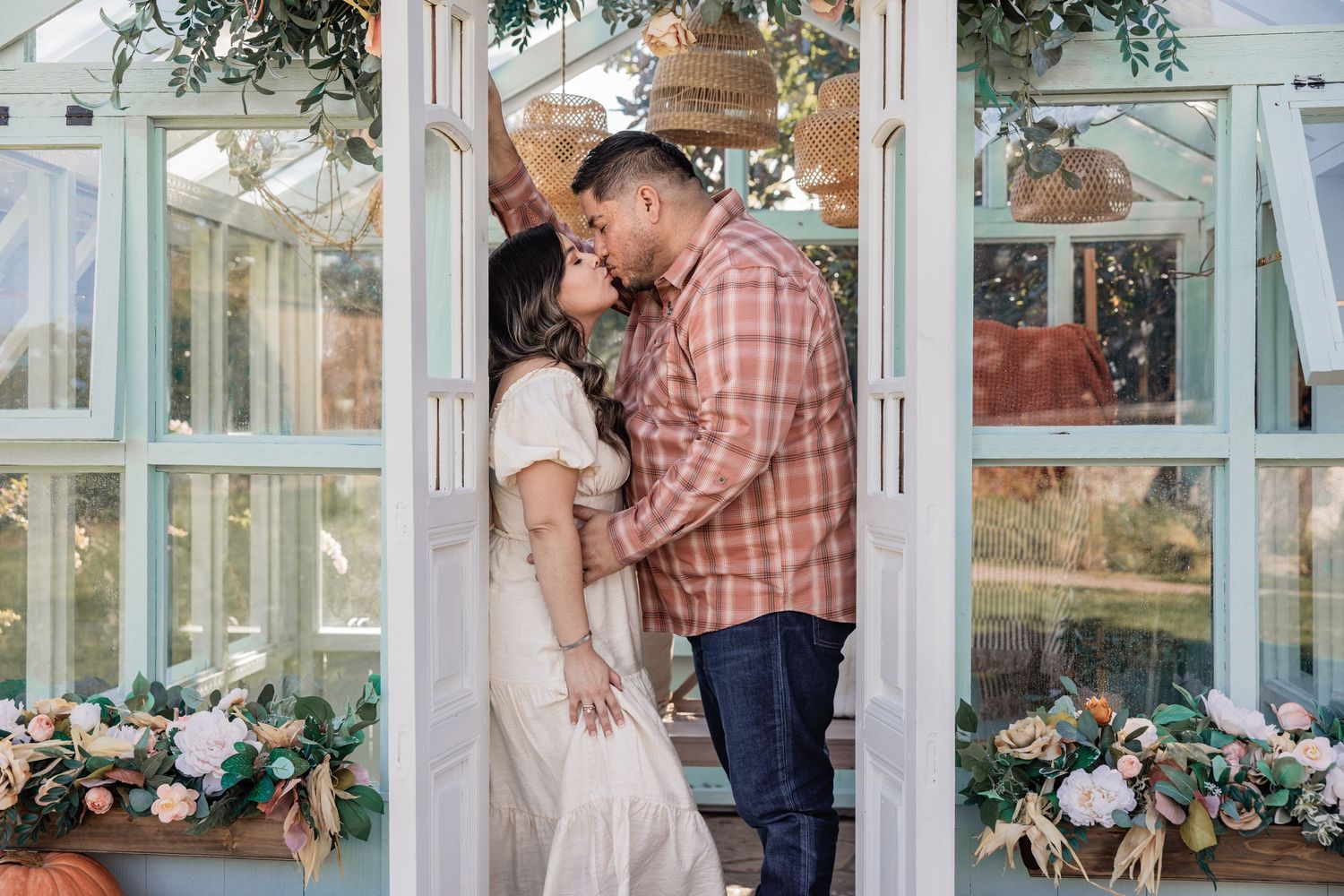 A couple shares a romantic kiss in a decorated doorway with floral arrangements and greenery.