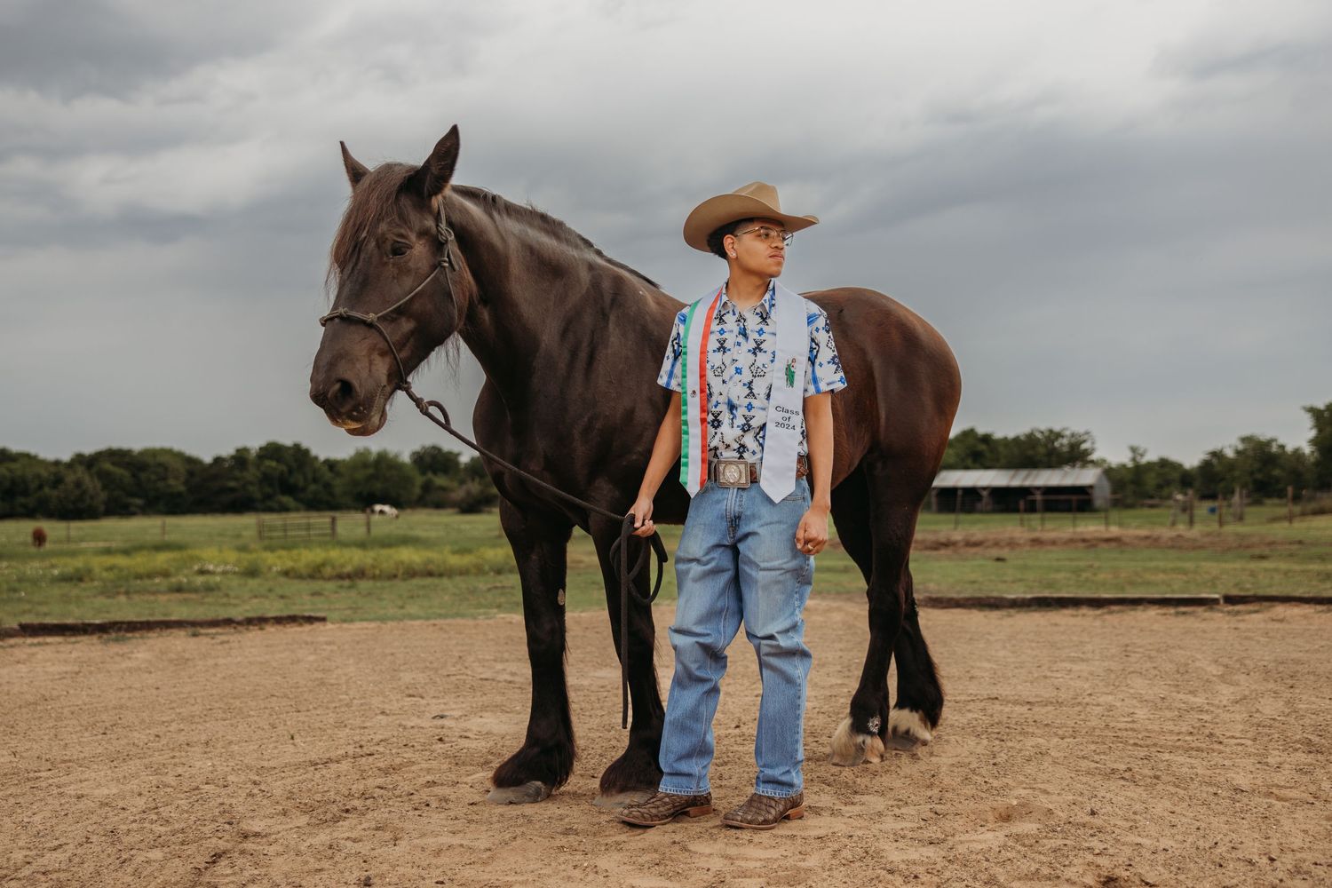 Teen boy in cowboy hat standing beside dark horse on ranch for senior photos.