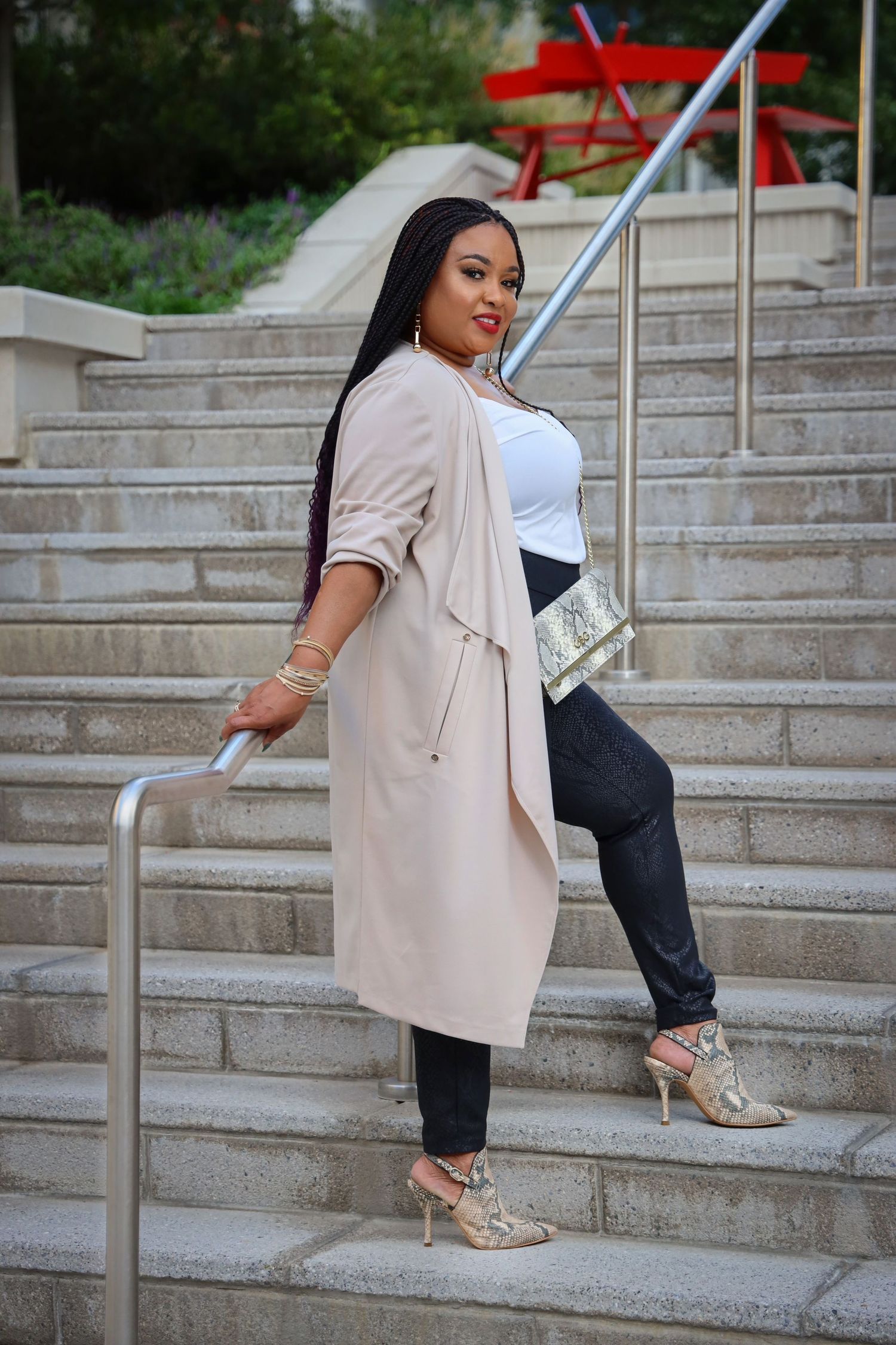 Stylish woman in a blush trench coat and snake print heels posing on outdoor stairs with a gold clutch.