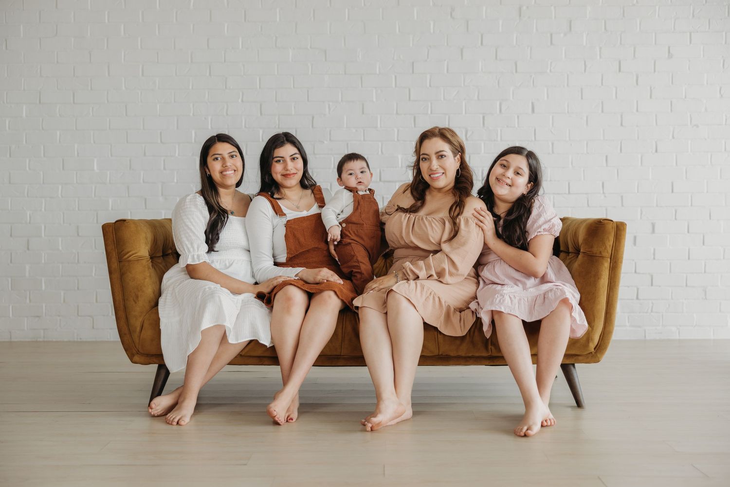 Five women sitting together on a mustard velvet sofa, one holding a baby, posing for a family portrait.