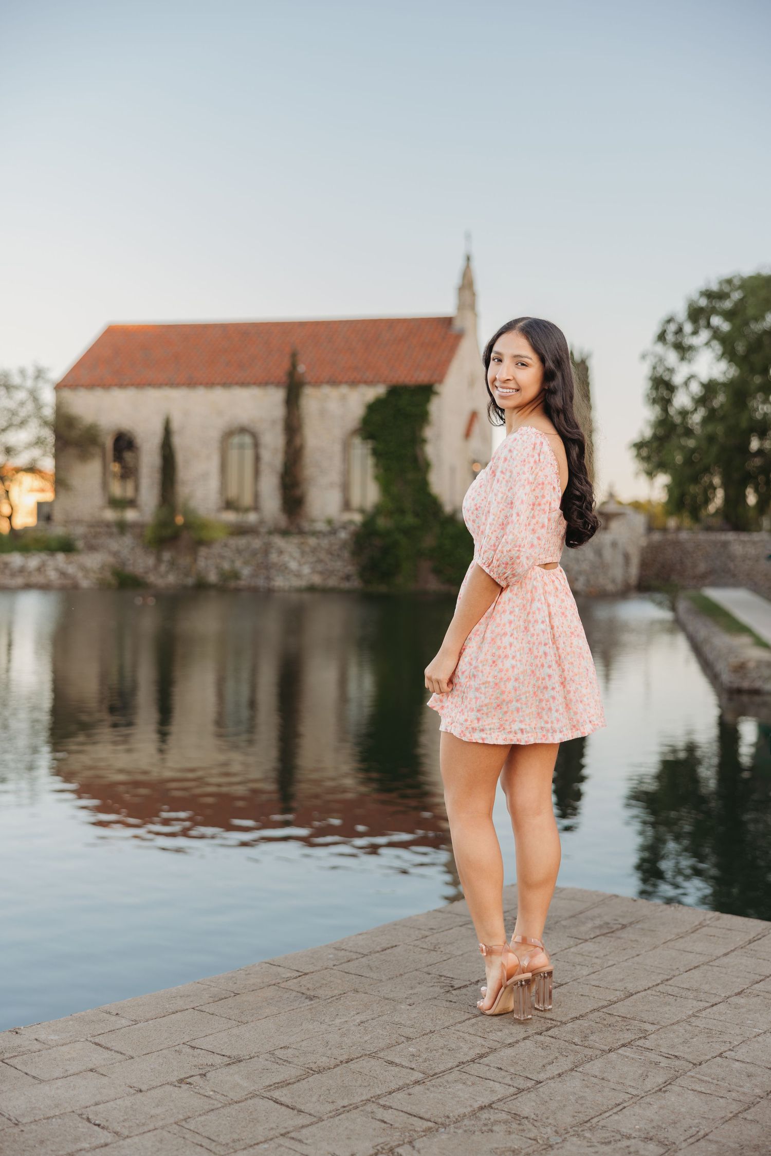Young woman in pink floral dress posing by reflective pond with historic chapel backdrop.
