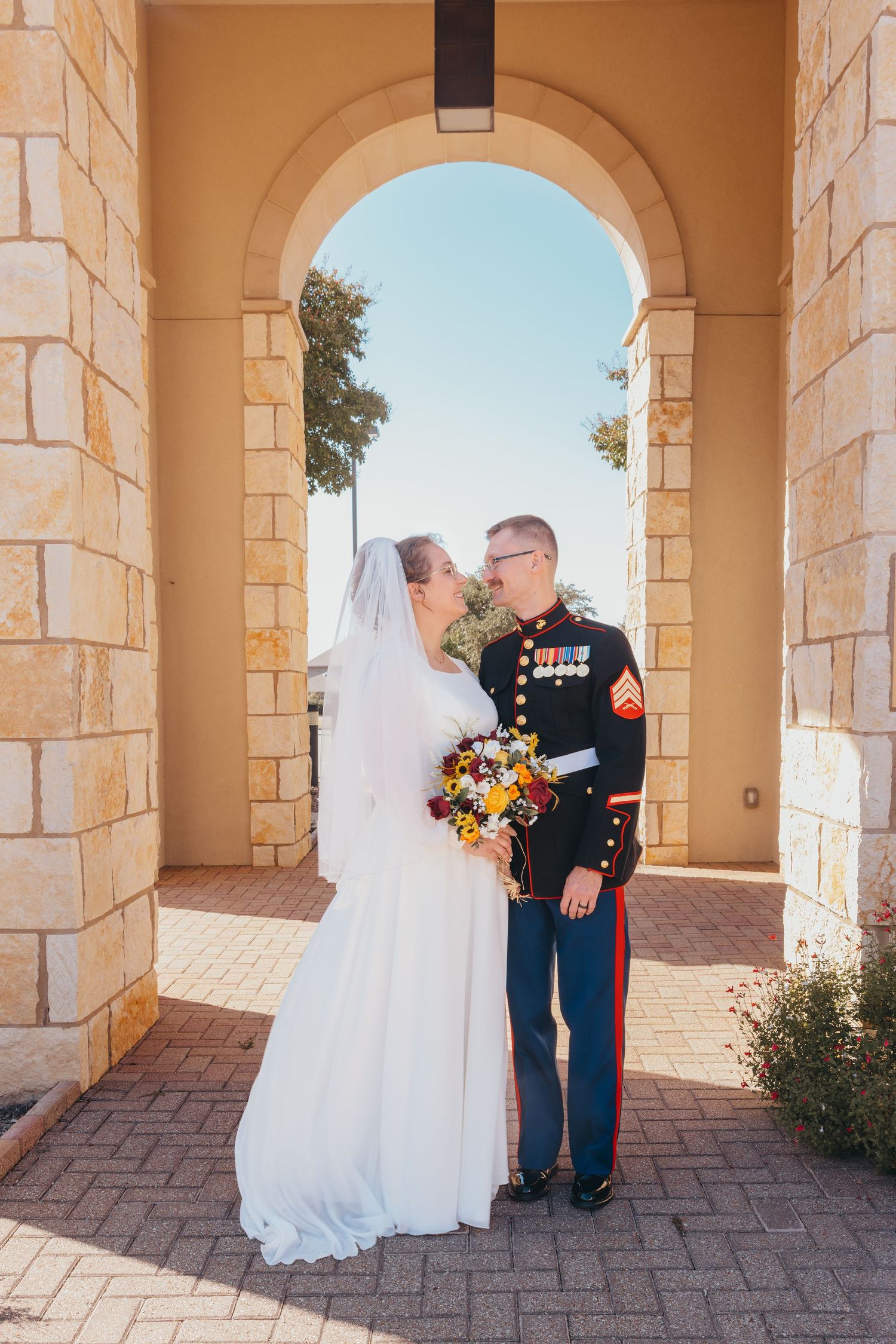 Military Marine groom in dress uniform and bride holding colorful bouquet posing under a stone arch.
