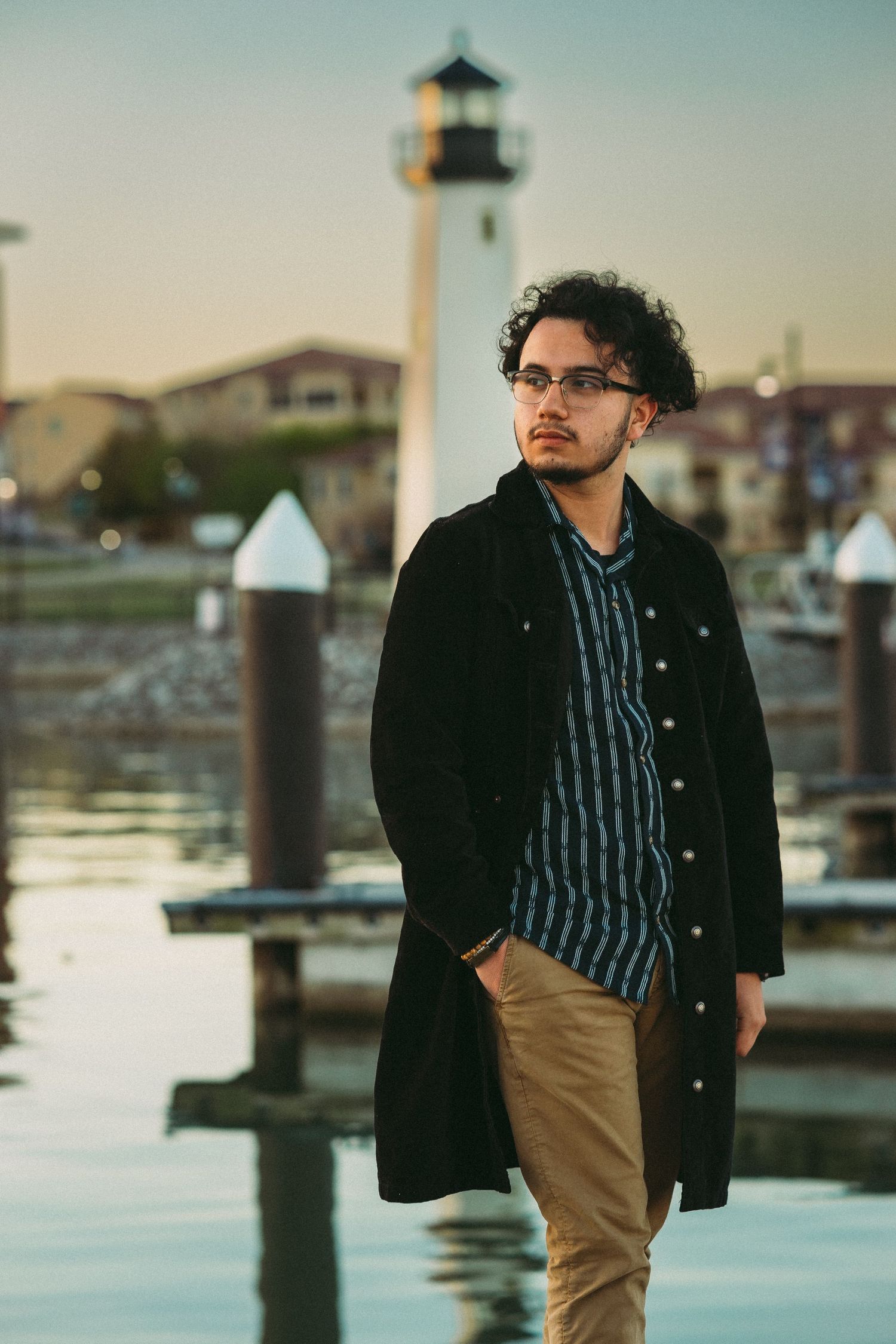 Young man in black coat and glasses stands near waterfront lighthouse at dusk.