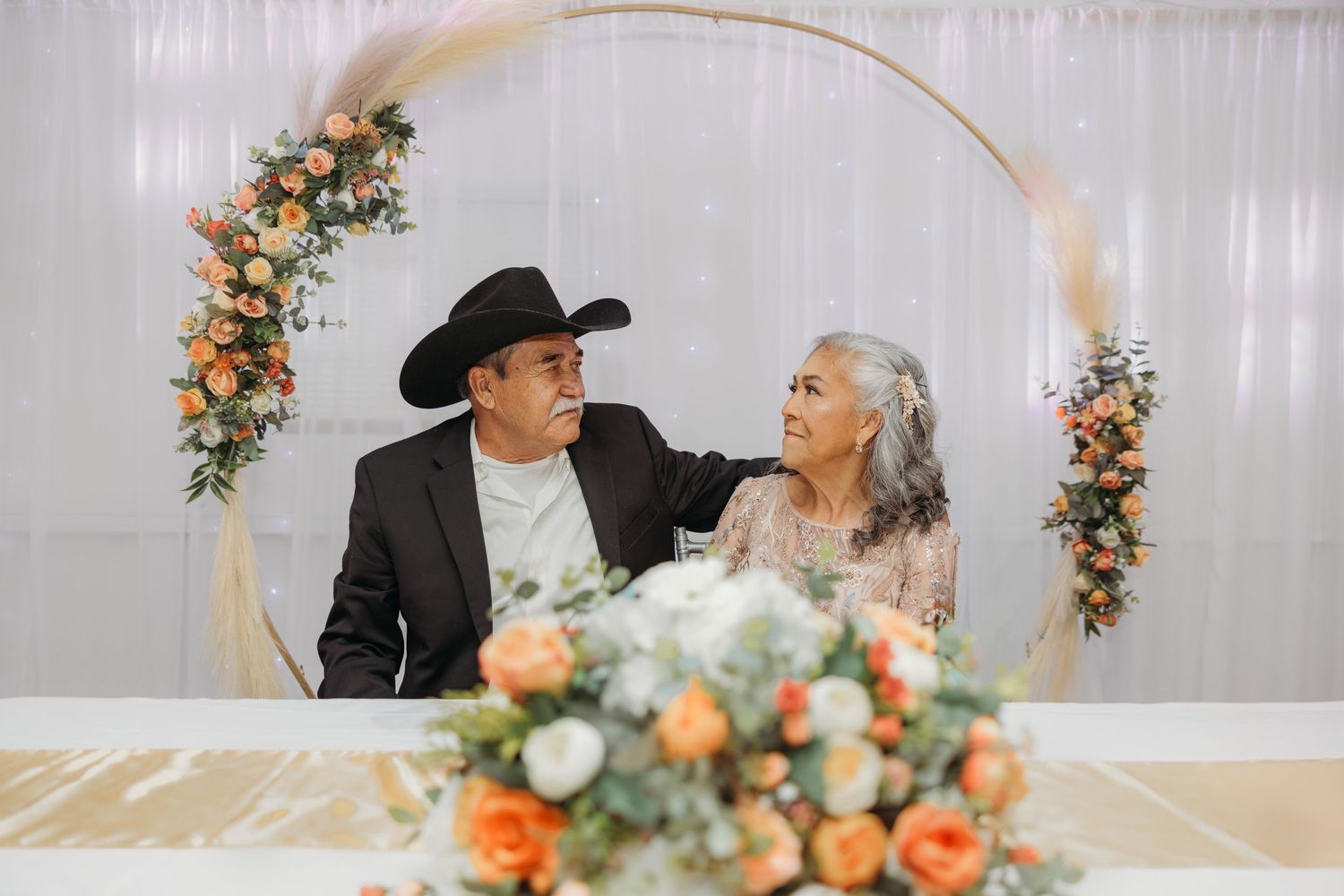 Couple seated at sweetheart table before circular floral arch at 50th anniversary celebration.