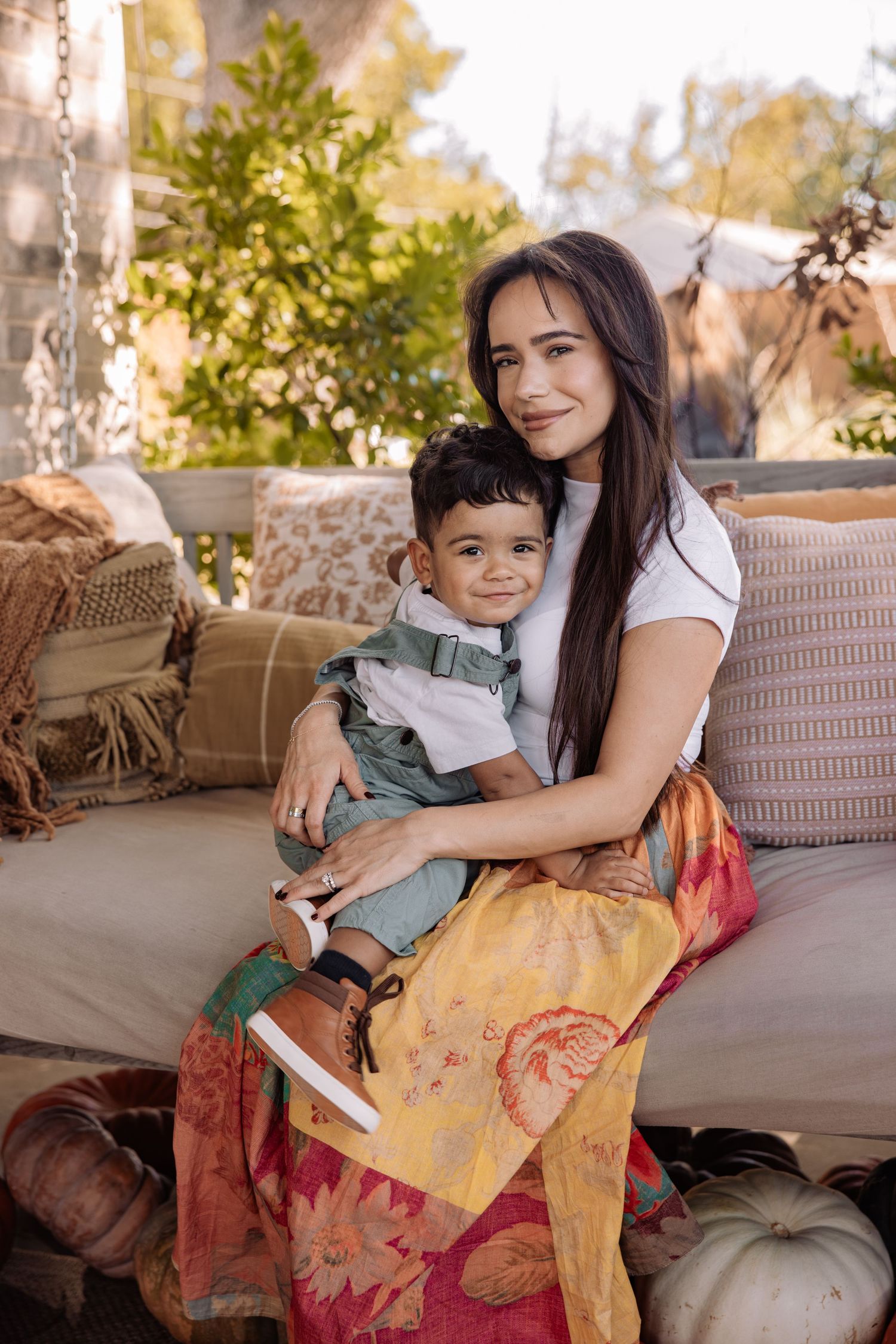 A young mother sits on a porch swing holding her toddler son, surrounded by autumn decor.