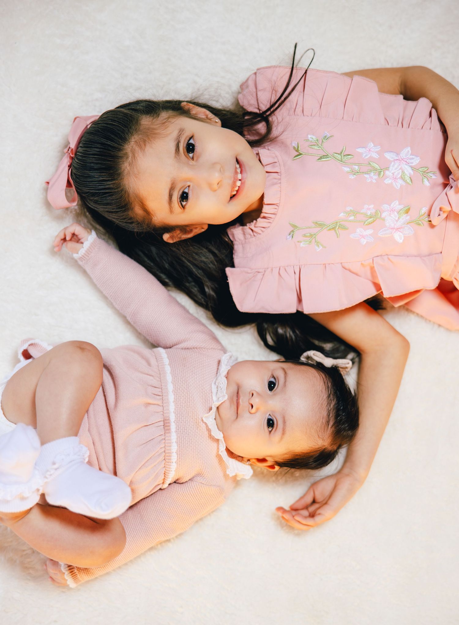 Two young sisters in matching pink ruffle dresses lying on a white carpet, smiling for a portrait photo.