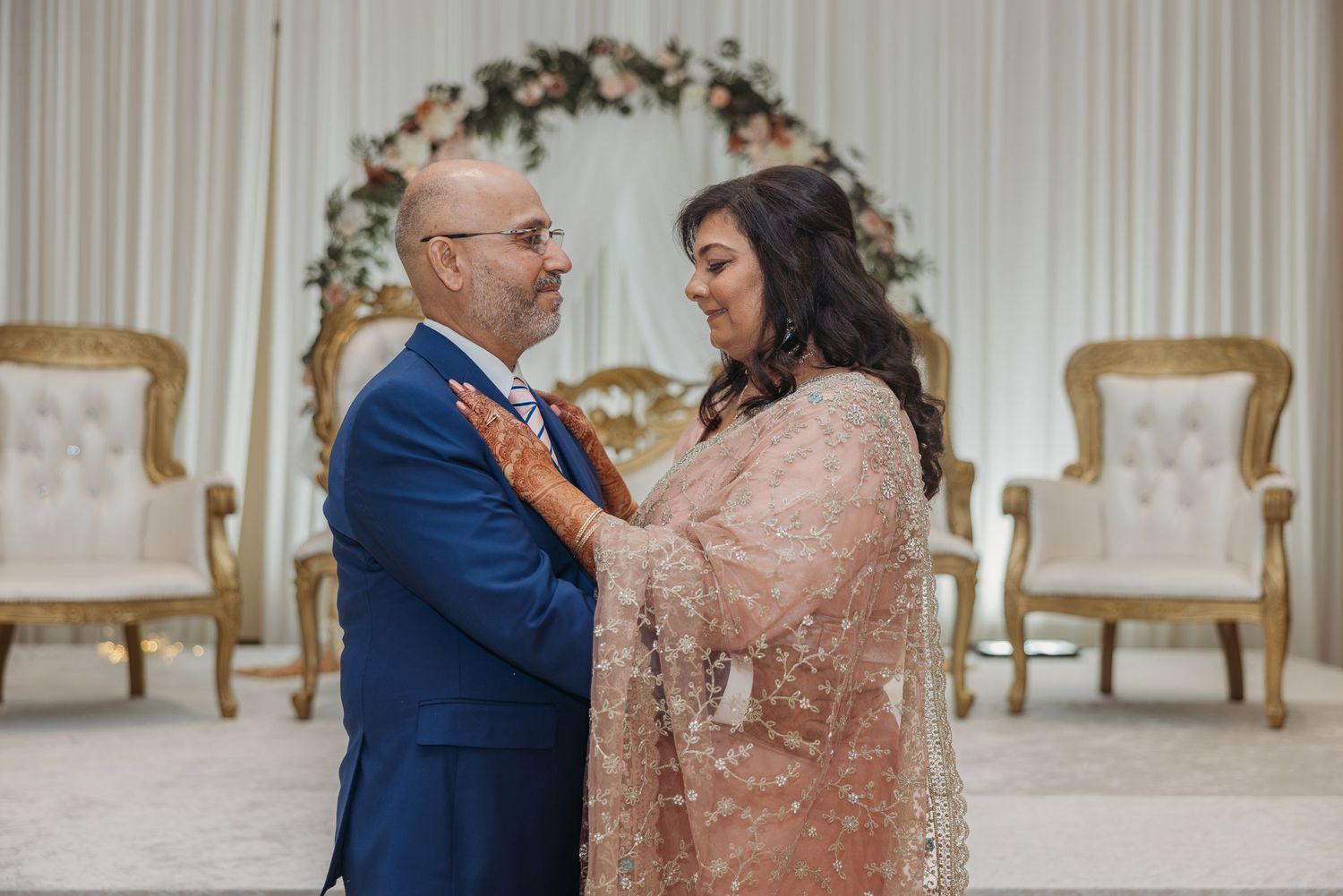 Couple in wedding attire smile at each other before floral arch with gold chairs.