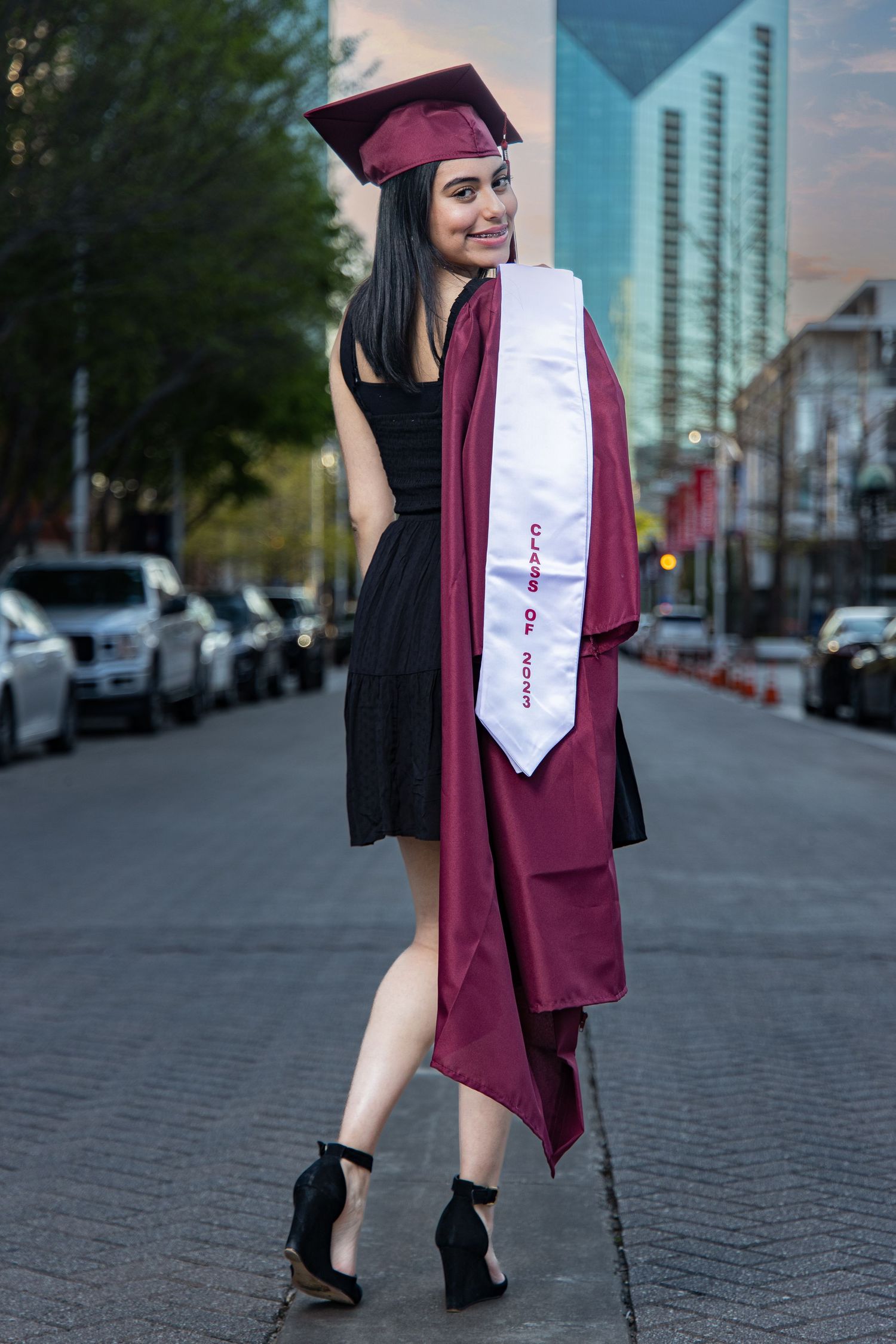 Female graduate in maroon cap and gown holding stole while posing on city street at dusk.