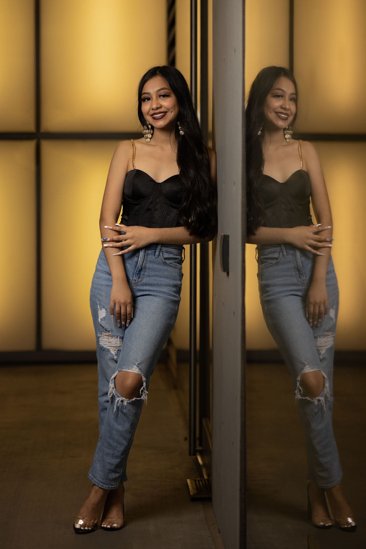 Smiling woman in black top and ripped jeans poses beside mirror reflection in golden light.