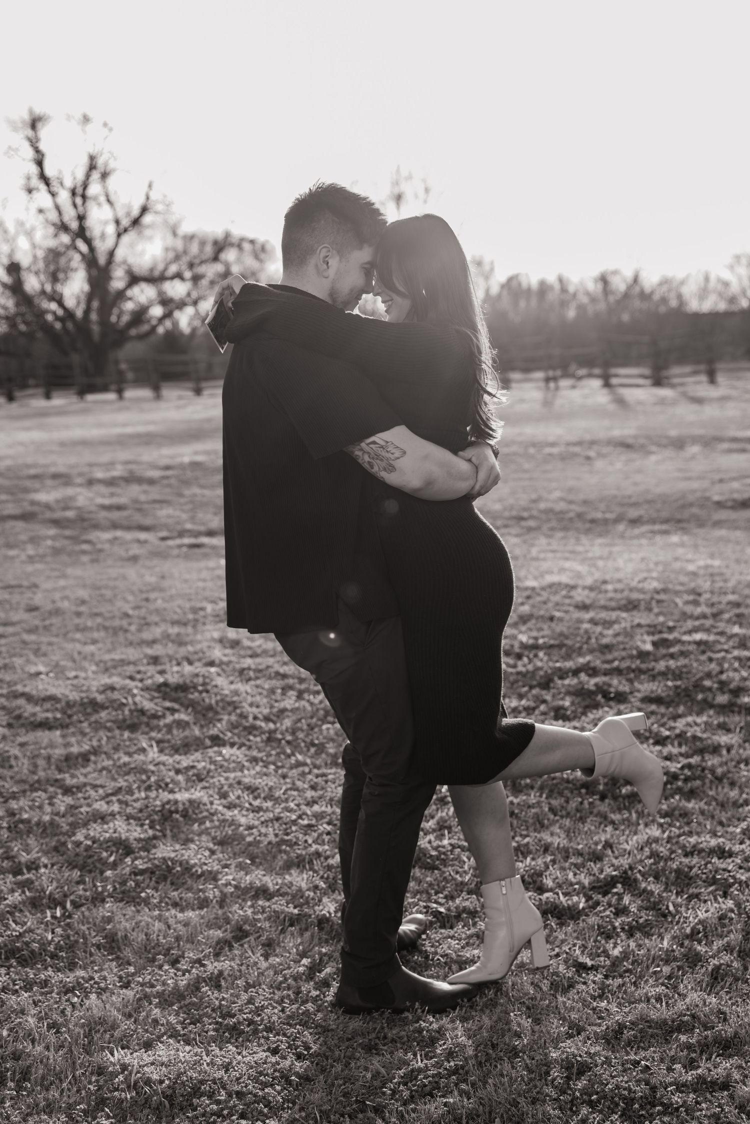 Black and white romantic photo of couple embracing and kissing in an open field at sunset.